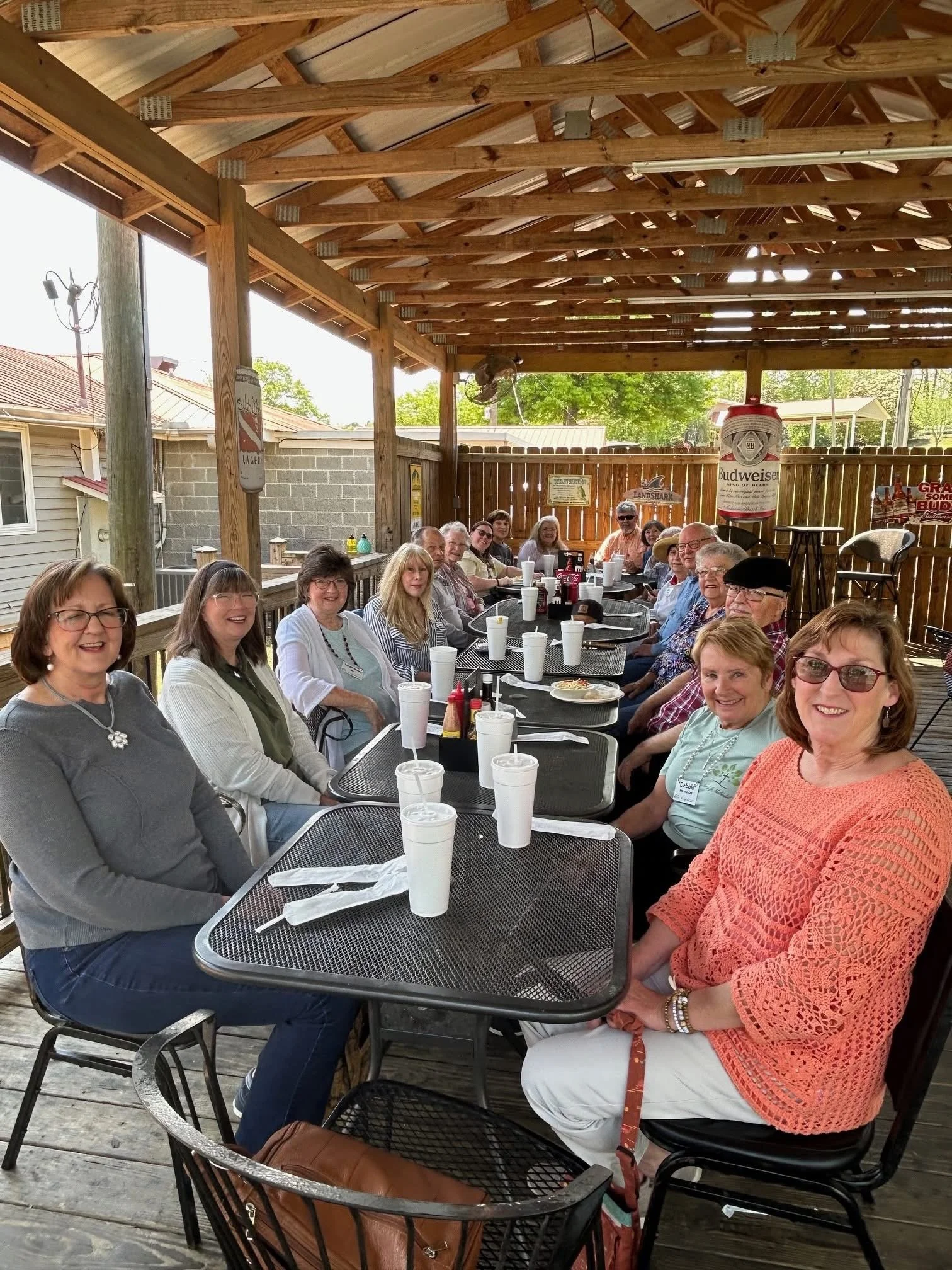 Group of women and men sitting at a long outdoor table under a wooden pavilion, with drinks and food, smiling for the camera.