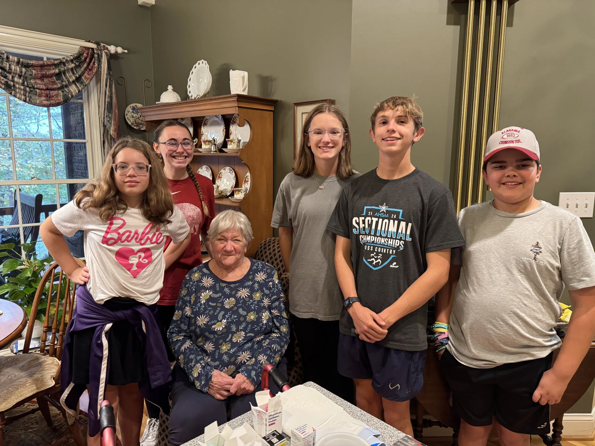 Six children and one elderly woman gathered together in a dining room, smiling at the camera. The woman is seated, surrounded by the kids standing behind her. The room features a green wall, a wooden china cabinet, a window with curtains, and a table