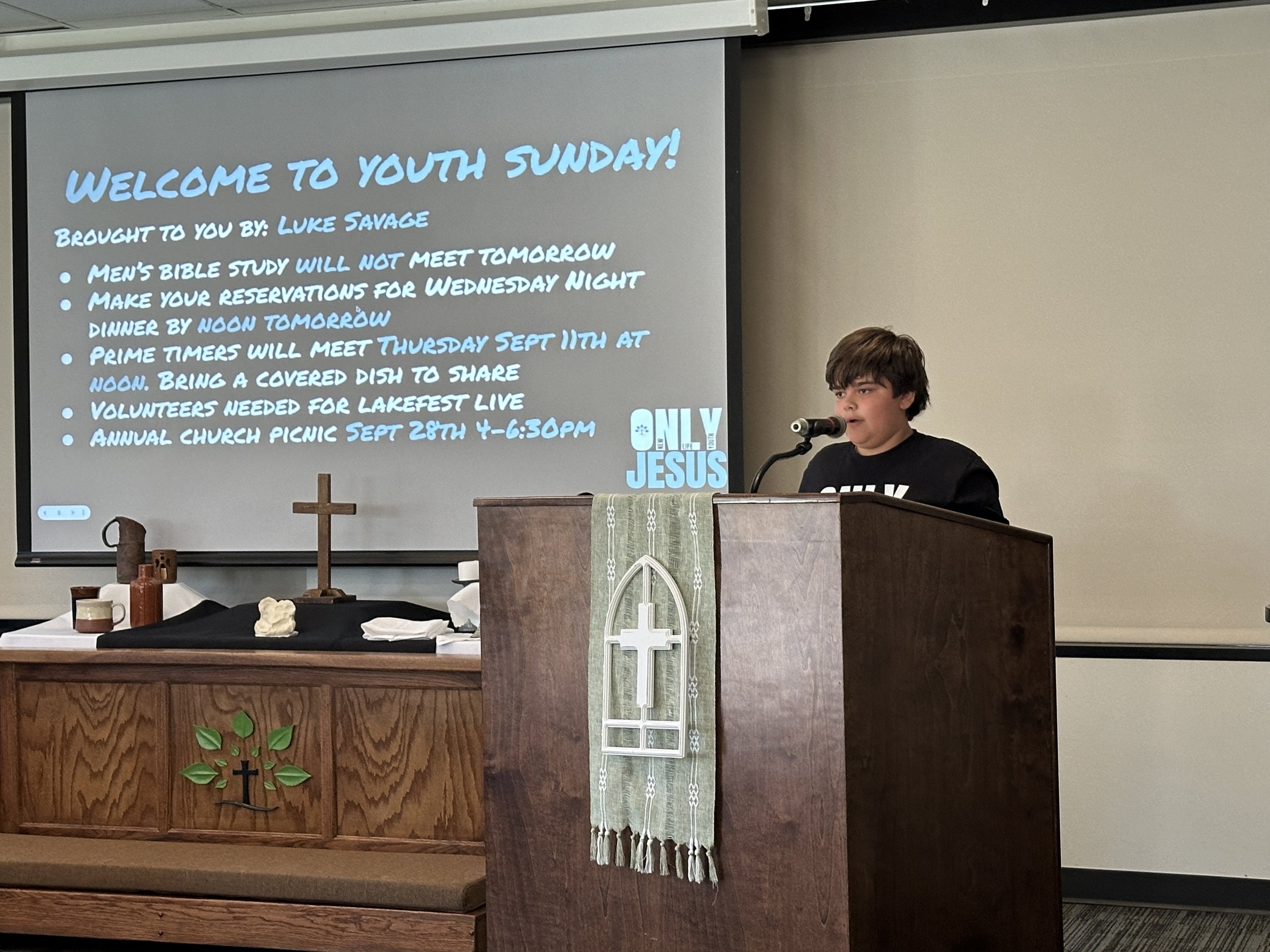 A young boy speaking at a church podium with a cross and a cloth with a church emblem. Behind him, a large screen displays a welcome message for Youth Sunday with details about upcoming church events and a note about only Jesus.