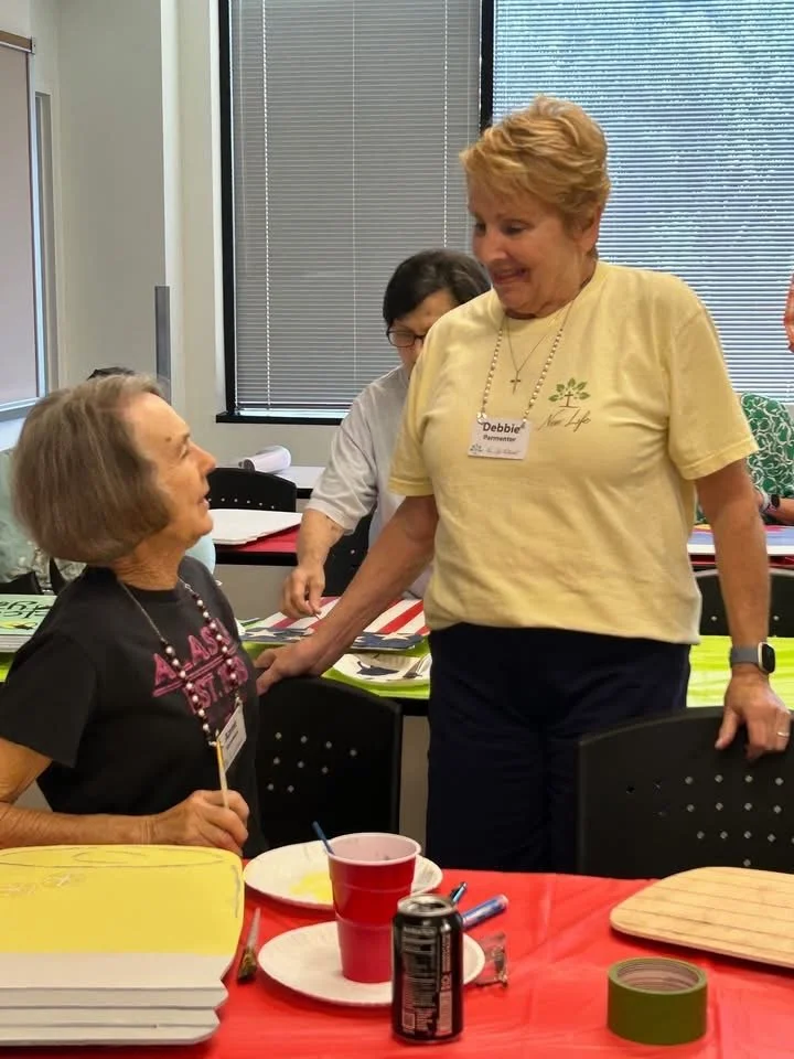 Two women engaging in conversation at a gathering or event, with one woman sitting and the other standing smiling, while another person in the background looks on. There are papers, a bowl, a soda can, and other items on the table.