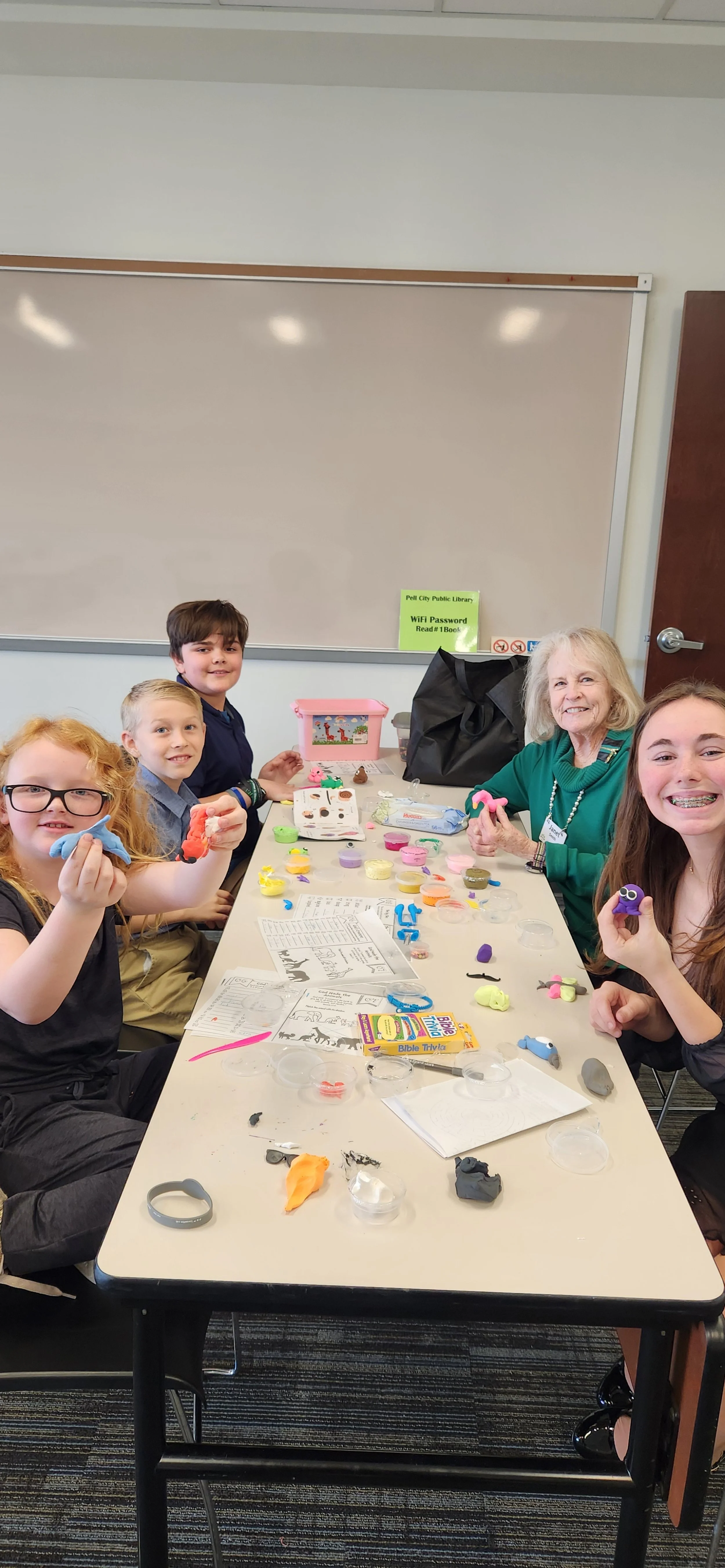 Children and an elderly woman making colorful clay crafts at a table in a classroom.