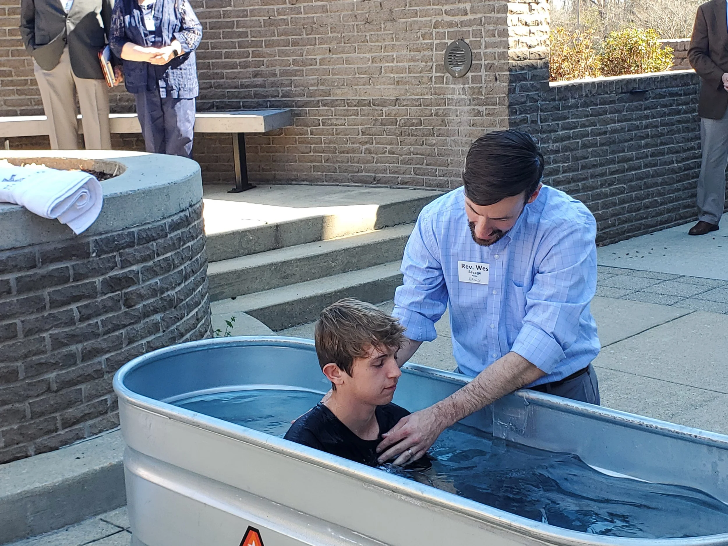 Young man submerged in water for baptism at outdoor ceremony, with minister assisting, surrounded by brick walls and other attendees in formal attire.