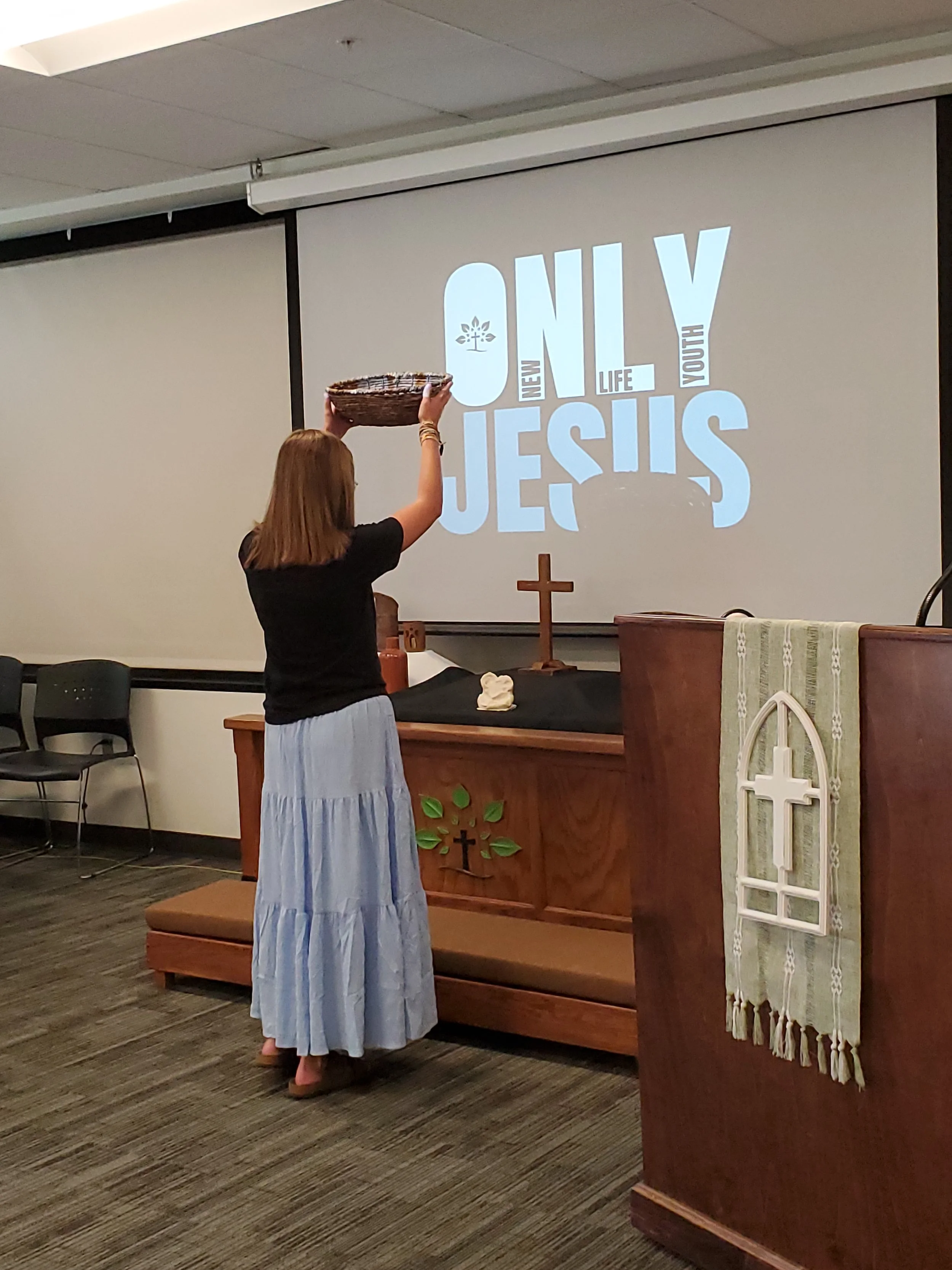 A woman with red hair wearing a black top and a long light blue skirt is placing a basket on a table in front of a church altar during a religious service.