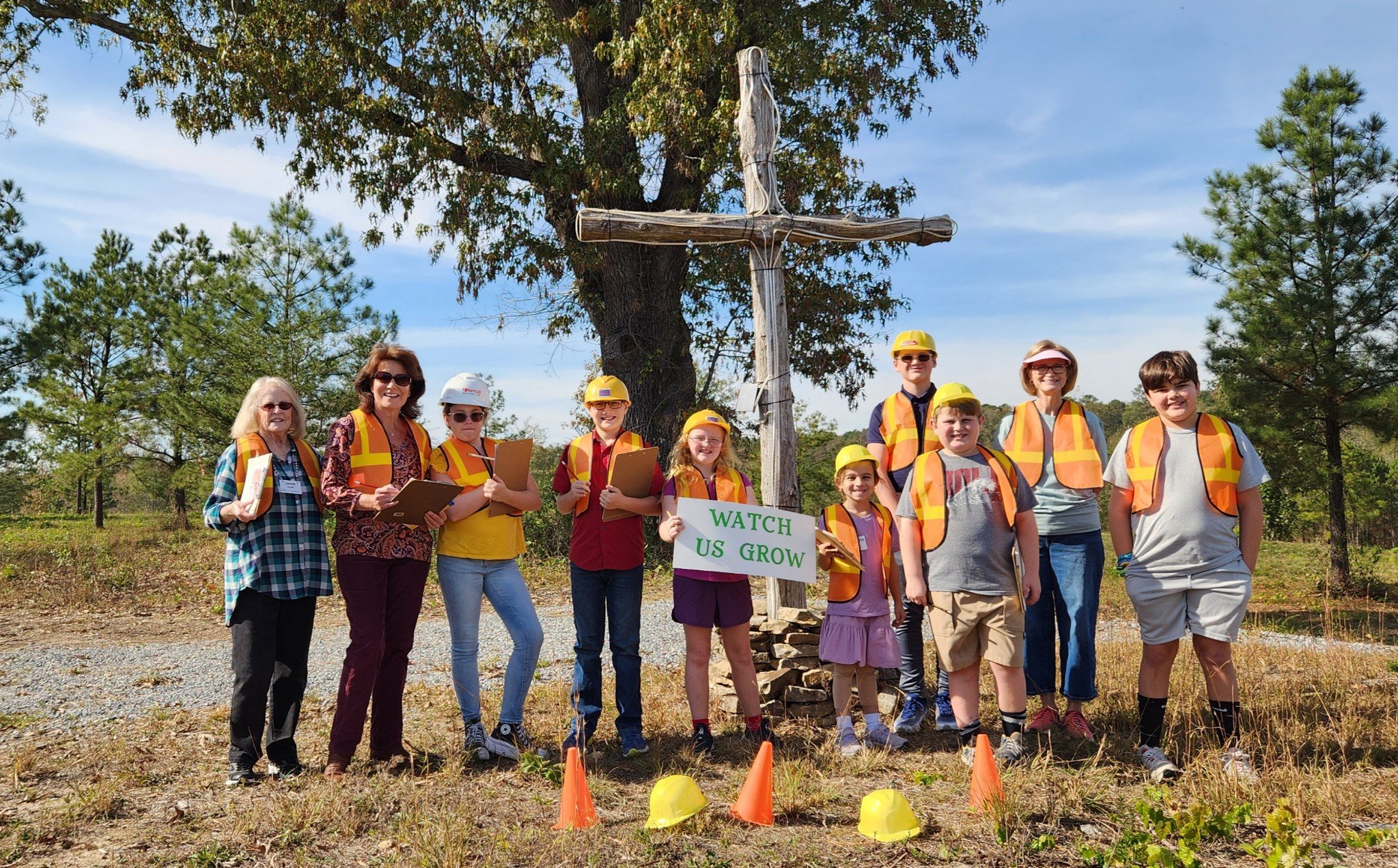 Group of children and adults in safety vests and helmets standing outdoors under a tree, holding clipboards and a sign that says 'Watch Us Grow', with a wooden cross on a small mound, surrounded by trees and cones.