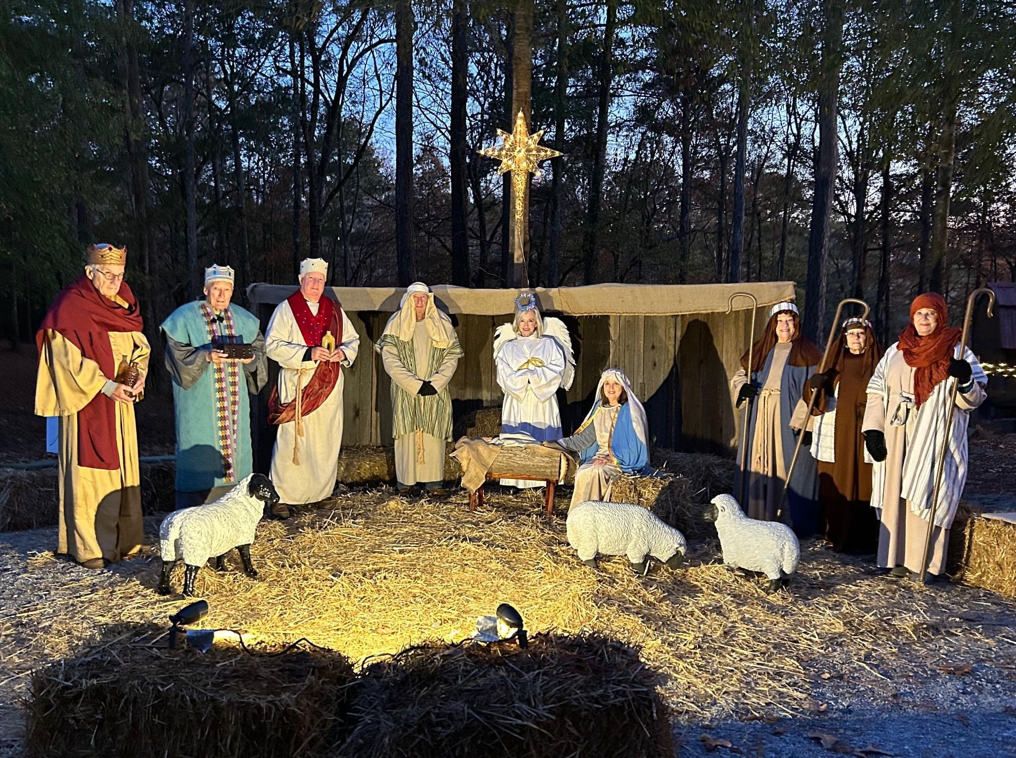 Nativity scene with figures of Mary, Joseph, baby Jesus, the three wise men, shepherds, and angels, set outdoors on a stage with hay and straw, illuminated by ground lights, with a star at the top, against a wooded background.