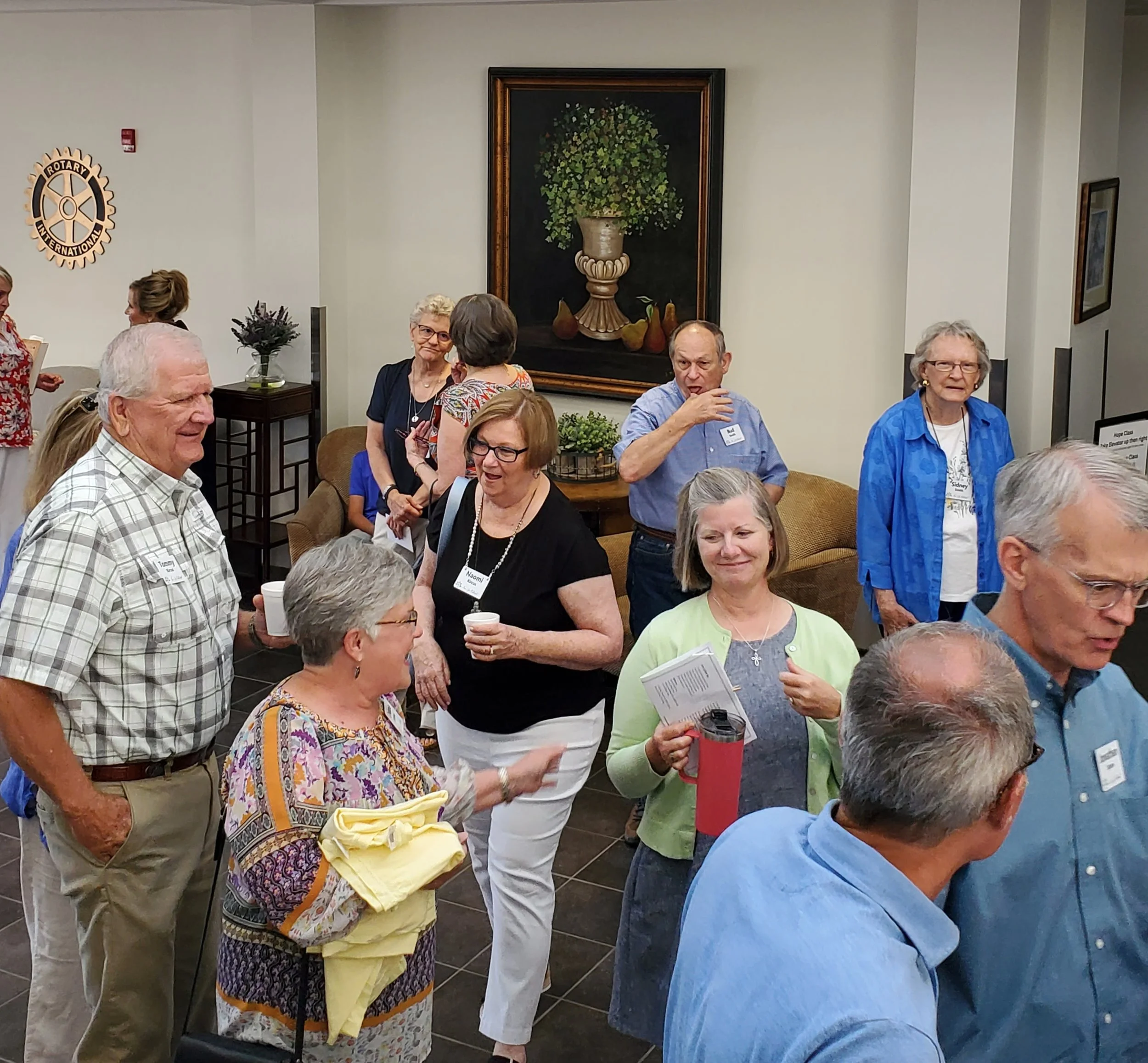A group of elderly people socializing at an indoor event, with some holding drinks or papers, in a room decorated with framed artwork and a floral arrangement.