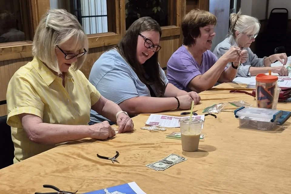 Four women sitting around a wooden table, engaged in a craft activity, with various supplies and a drink in a clear plastic cup with a straw.