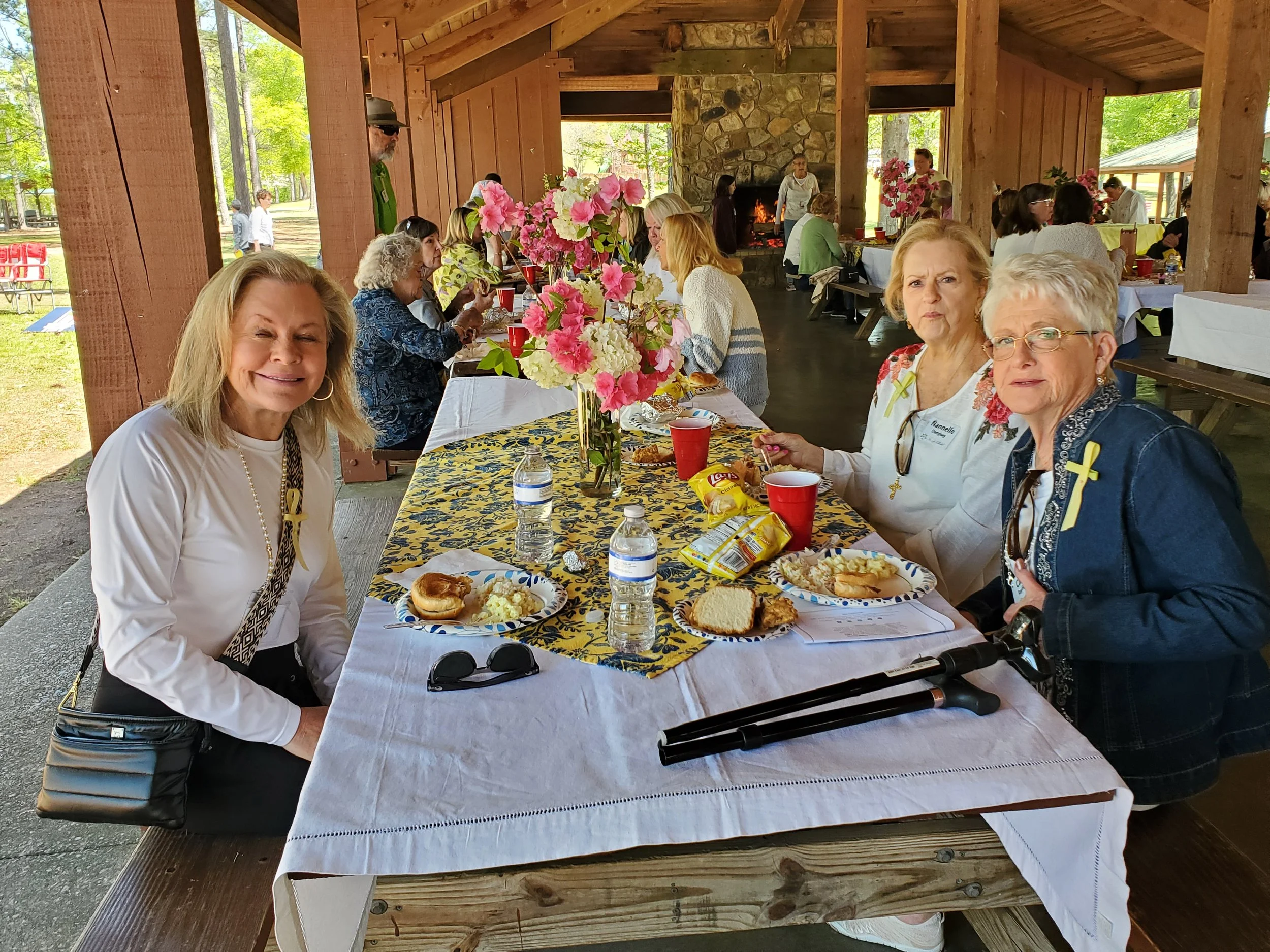 Group of women sitting at a decorated outdoor dining table with food and drinks, under a wooden pavilion with a fireplace, in a park with trees.