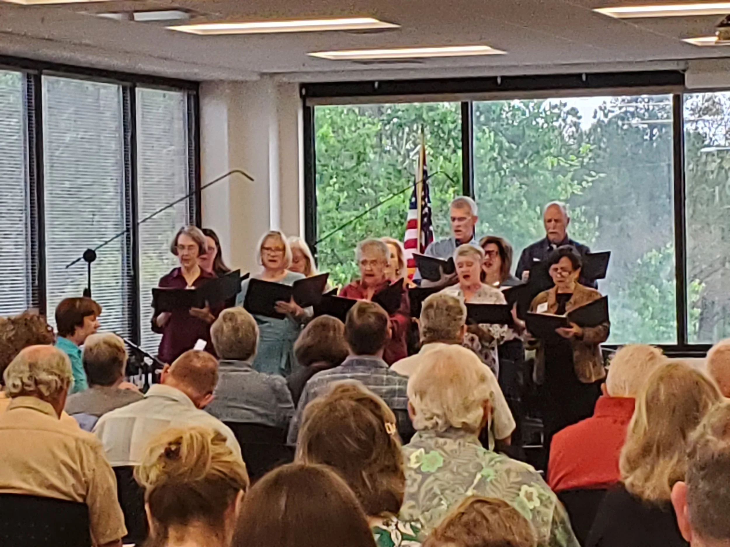 Group of elderly choir members singing at a community event in a room with large windows and an American flag, audience seated in front.