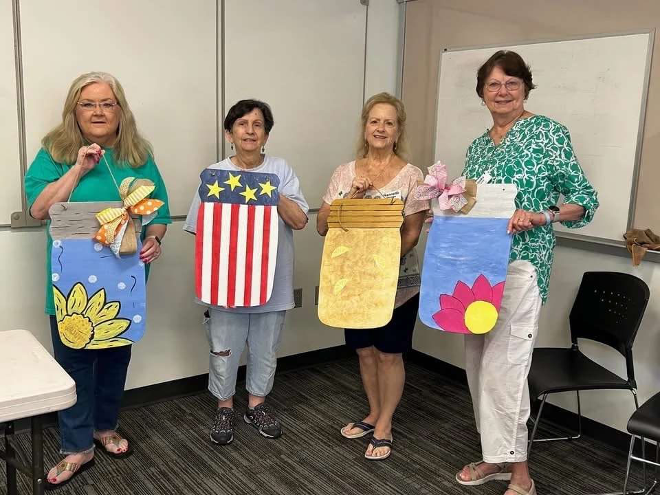 Four women holding large, painted paper bags with decorative bows, standing in a classroom.