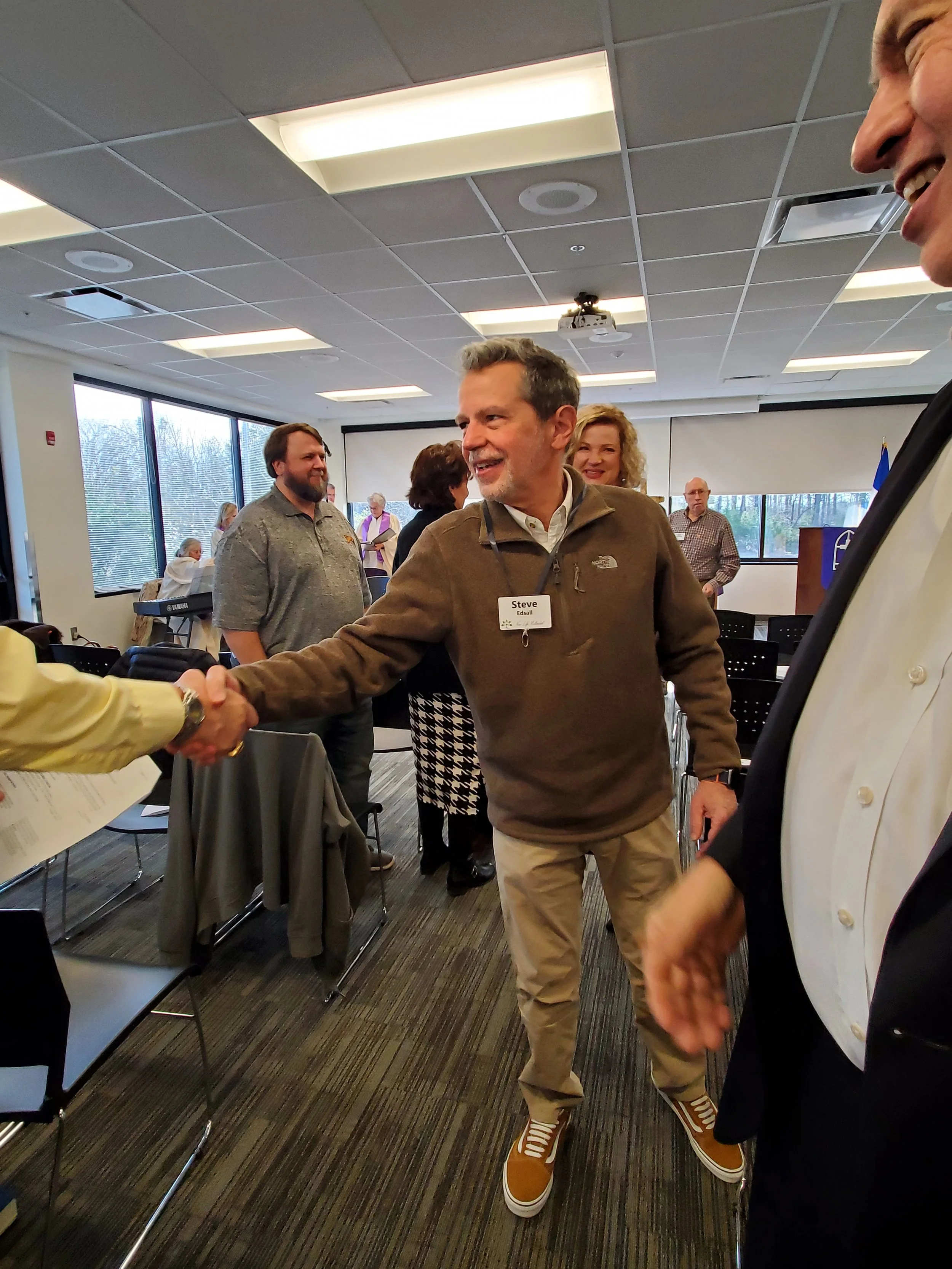 Man wearing a brown jacket and khaki pants shaking hands with a person in a yellow shirt in a conference room. Other people are standing and sitting in the background.