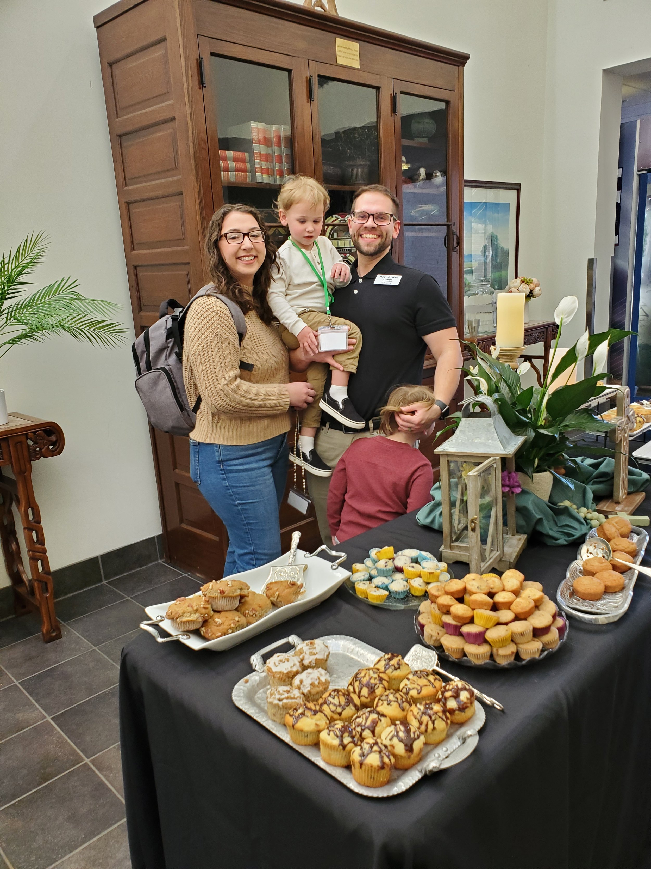 A family of four standing behind a table filled with various cupcakes and baked goods at a social event, with a wooden cabinet and framed picture in the background.