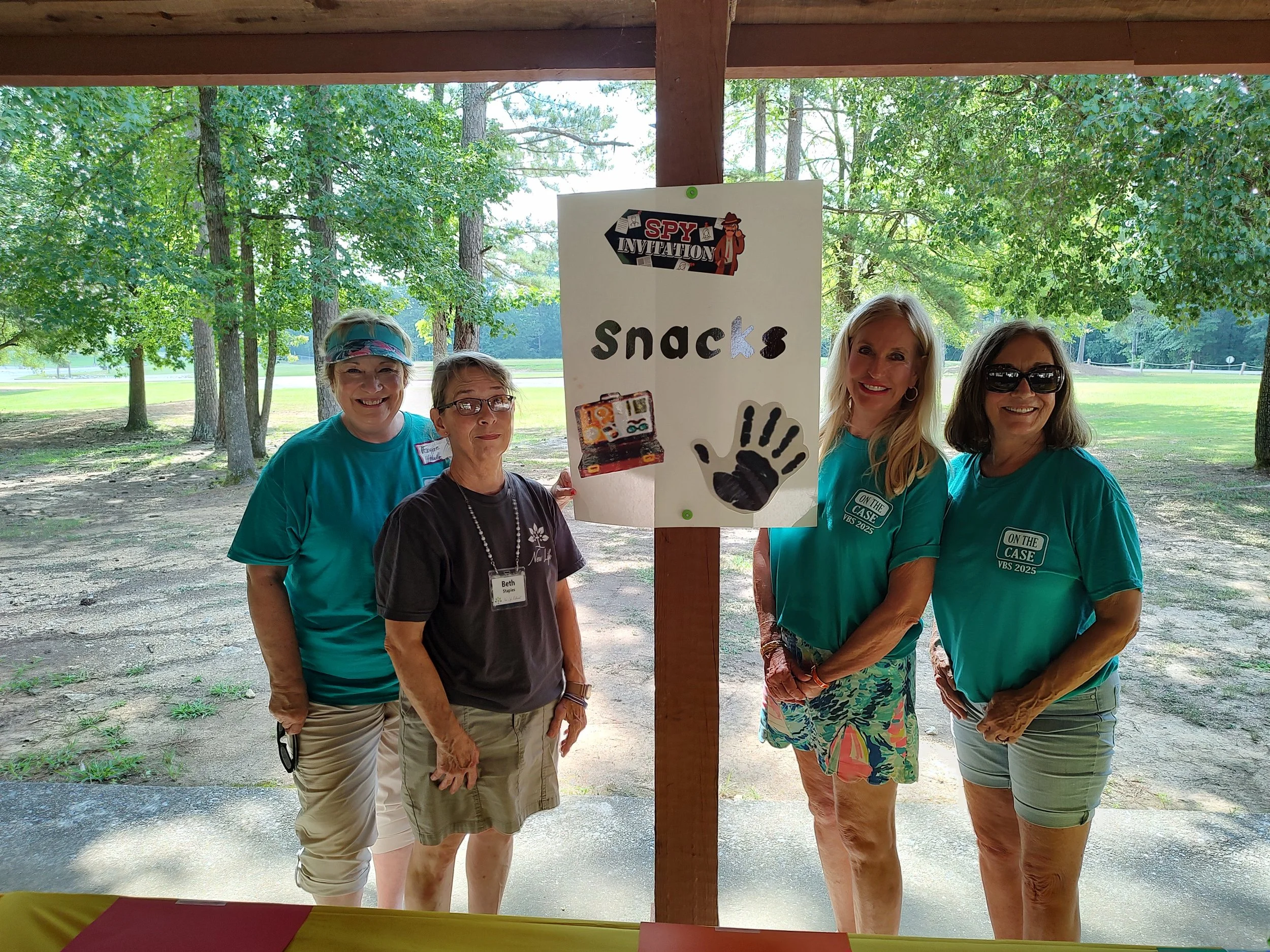Four women standing outdoors near a table with a sign that says 'Snacks' and depicts a hand, a candy bar, and a spy theme. The women are smiling and wearing casual summer clothing.