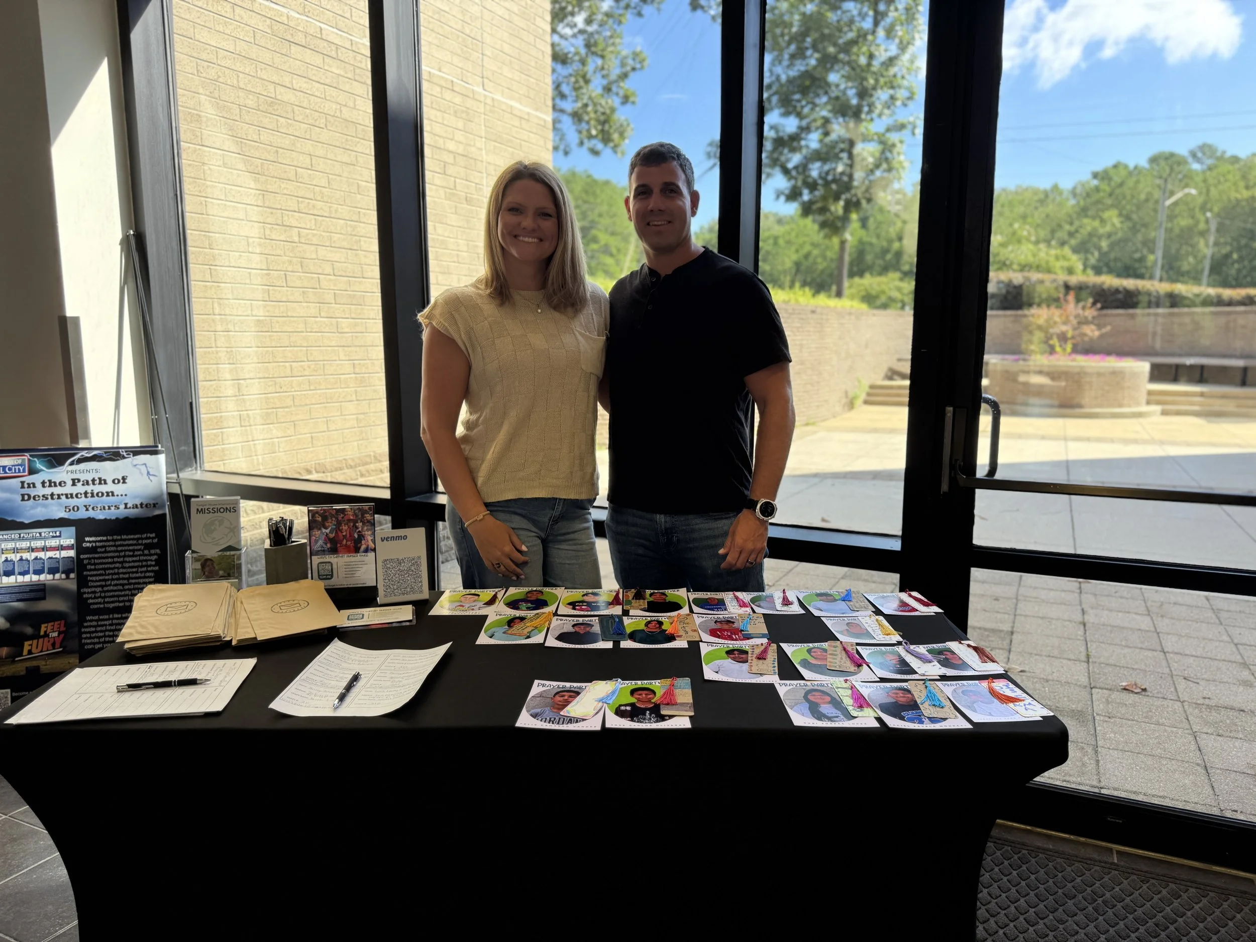 Two people standing side by side behind a table with pictures of children, papers, and signs inside a building with large windows and a view of a brick courtyard and trees outside.