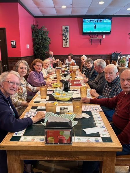 A group of elderly people sitting around a long table in a restaurant, with drinks and menus, pink walls, a wall-mounted TV showing a tennis match, and some decorations.
