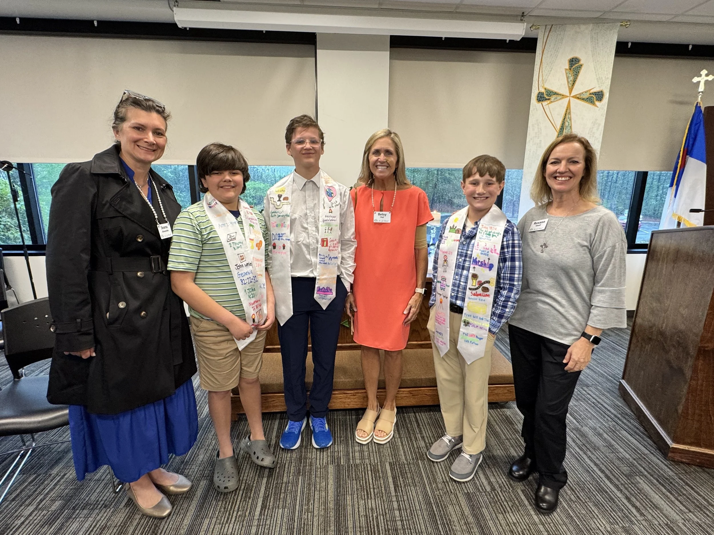 Group of six women and children standing inside a room, some wearing sashes with religious motifs, in front of a wooden pulpit, with flags and religious banners in the background.