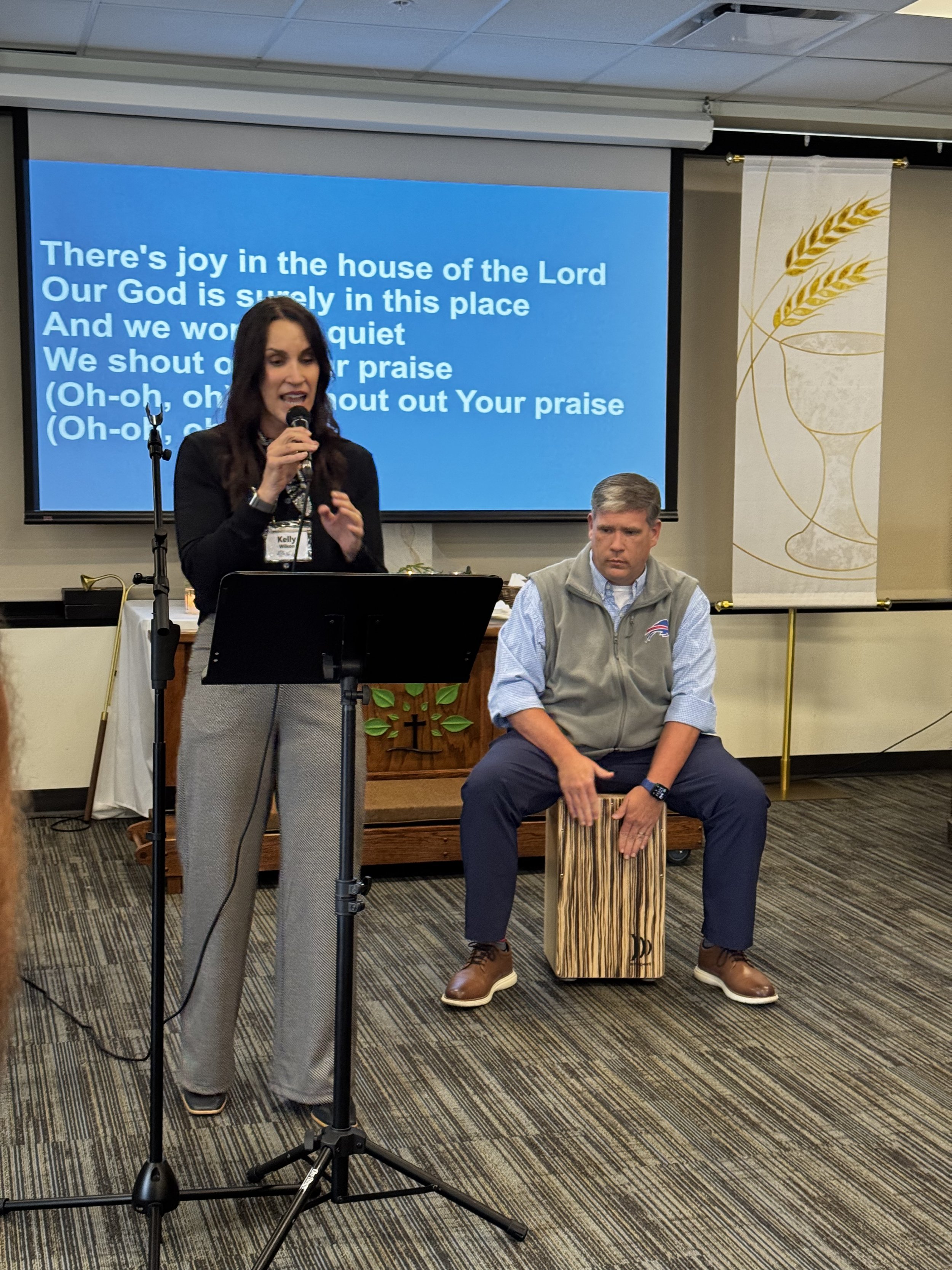 A woman singing into a microphone while a man plays a cajón drum in a church setting. There is a large screen behind them displaying lyrics of a hymn, and a church banner with wheat design is visible on the right.