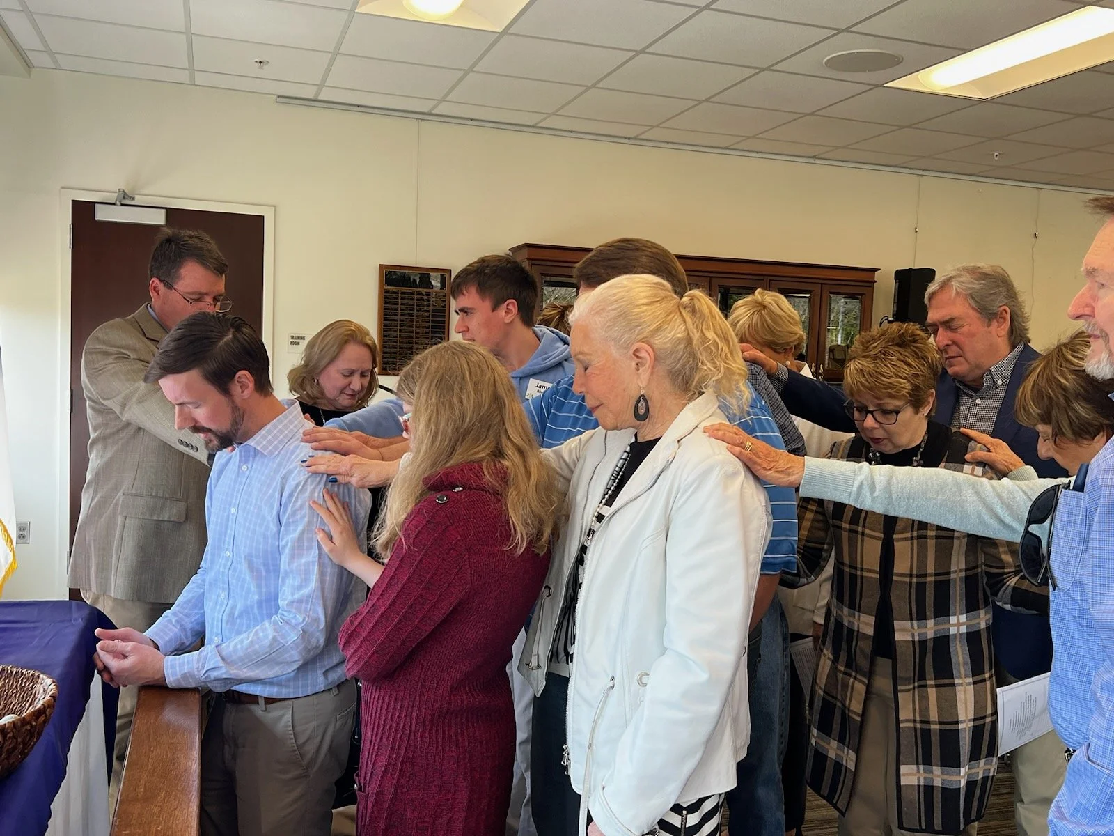 Group of people praying or blessing each other with hands placed on shoulders in an indoor setting.