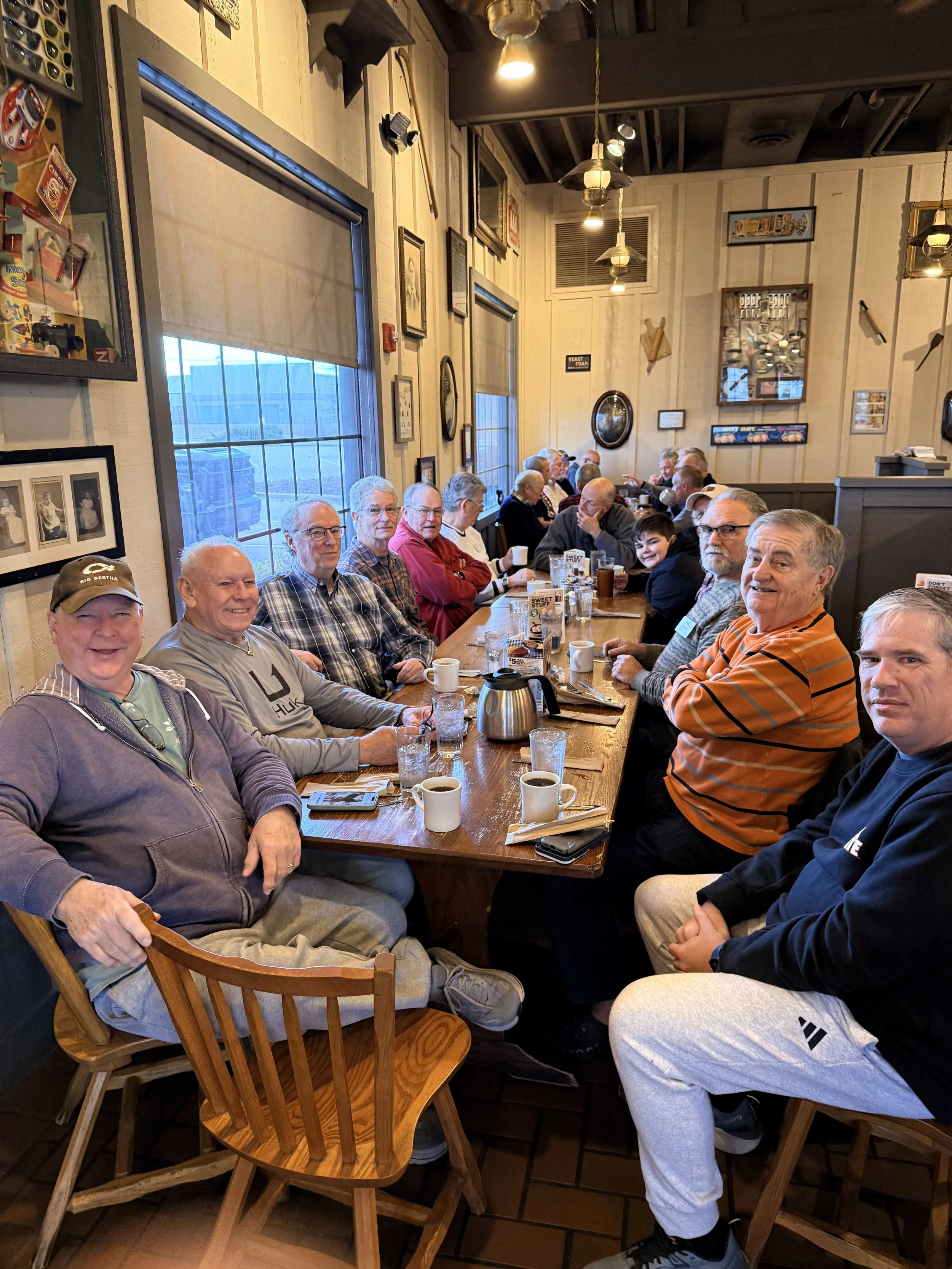 A group of elderly men and a young boy sitting around a long wooden table in a cozy restaurant with wooden walls decorated with framed pictures and various memorabilia. They are enjoying coffee and conversation.