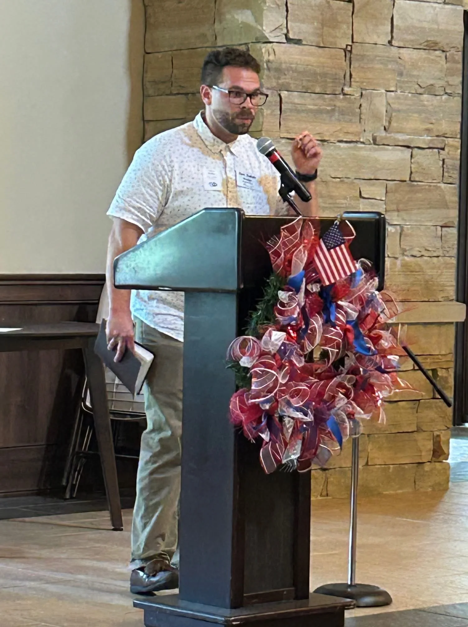 Man with glasses and scruffy beard, wearing a short-sleeved, collared shirt, standing at a podium decorated with a patriotic wreath of red, white, and blue ribbons, small American flags, and a blue bow, giving a speech into a microphone.