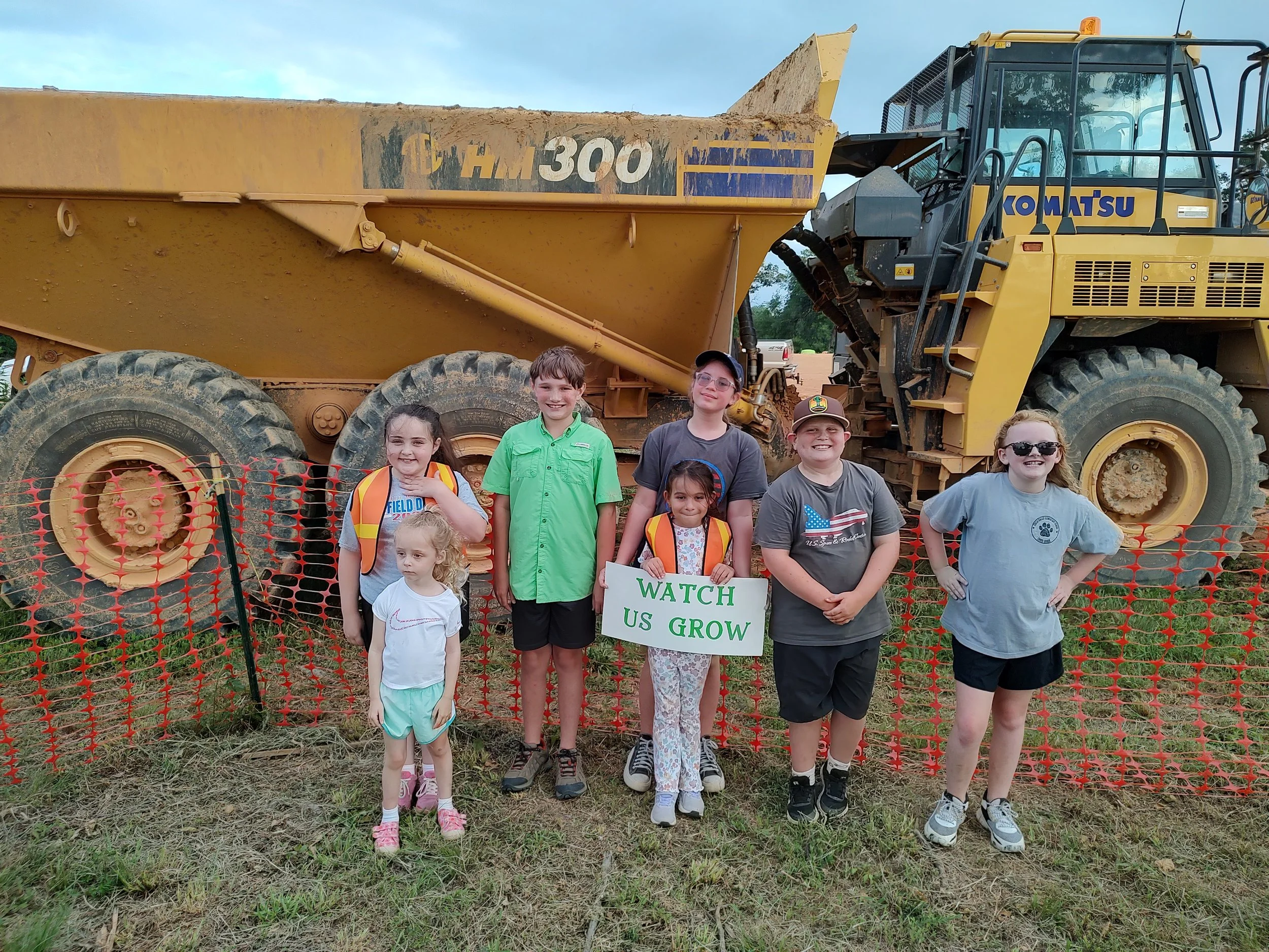 Group of seven children standing in front of a large construction dump truck, holding a sign that says 'WATCH US GROW,' at an outdoor event or farm.