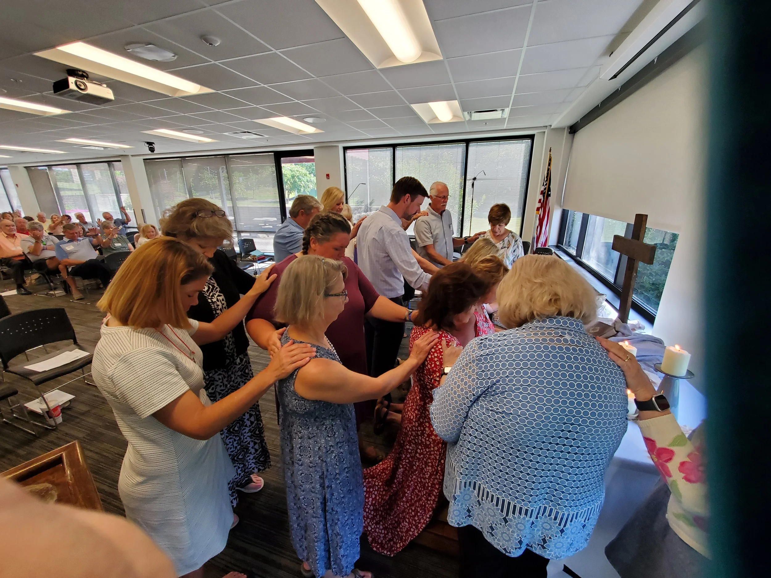 People praying at a religious service in a church or community center. They are kneeling or standing with heads bowed, facing a table with lit candles, a cross, and a small altar against a window.