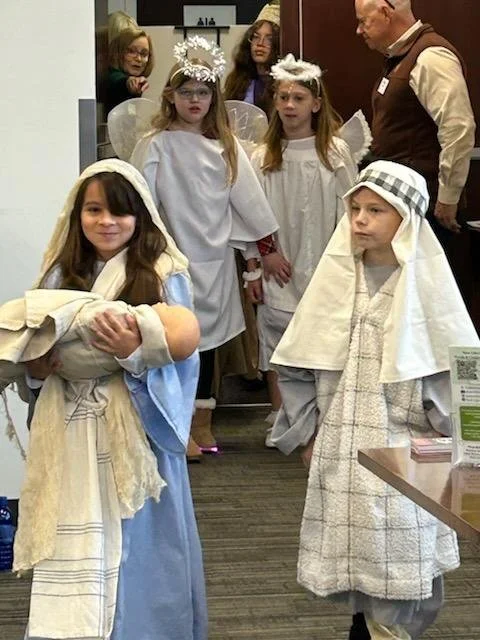 Children dressed as angels in a nativity scene, with a girl holding a doll wrapped in a blanket, and other kids and adults observing.