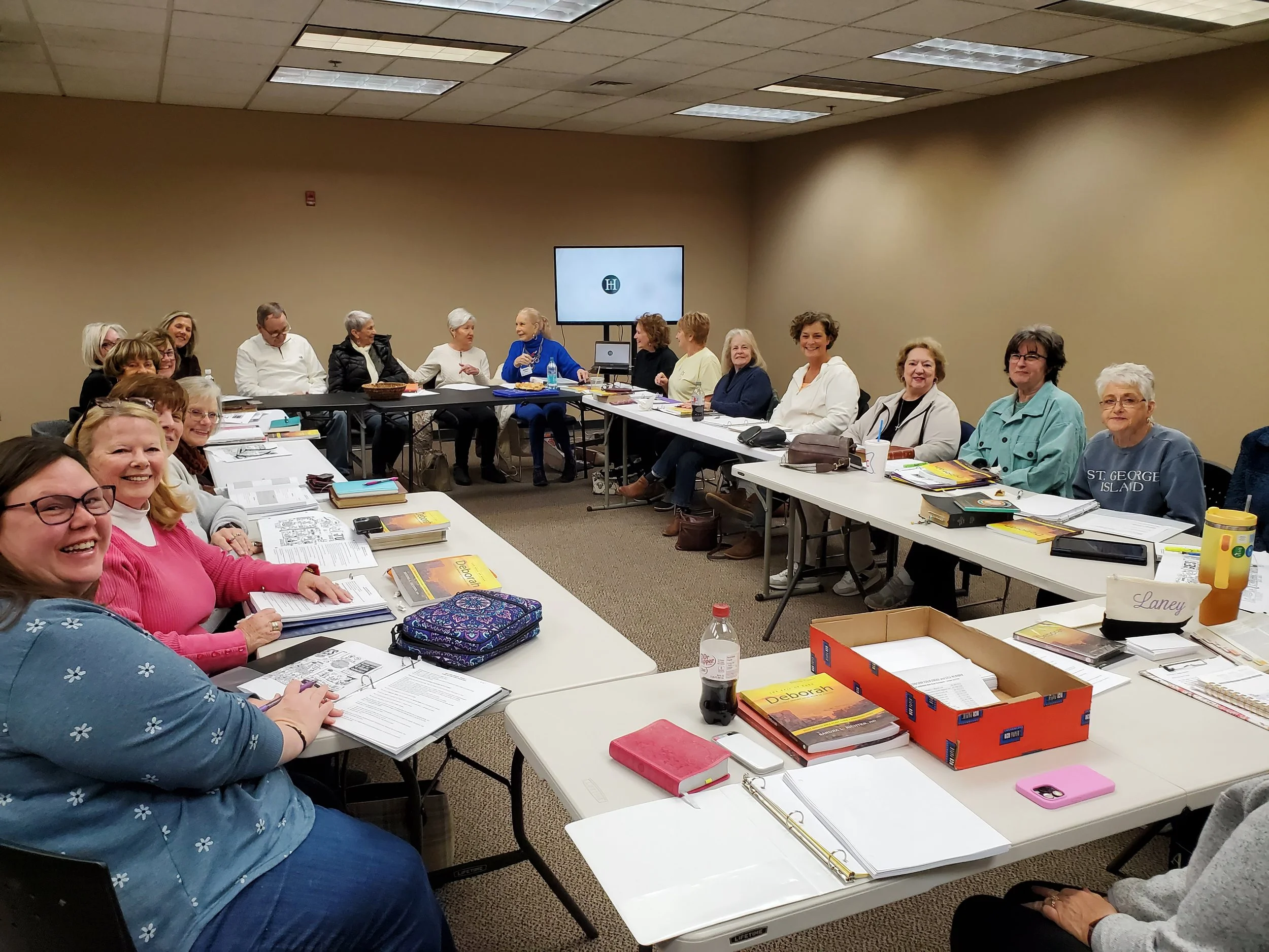 A group of women sitting around a U-shaped conference table in a meeting room, smiling and engaging. The table is cluttered with books, notebooks, papers, folders, water bottles, and electronic devices. A large monitor is on the beige wall at the bac
