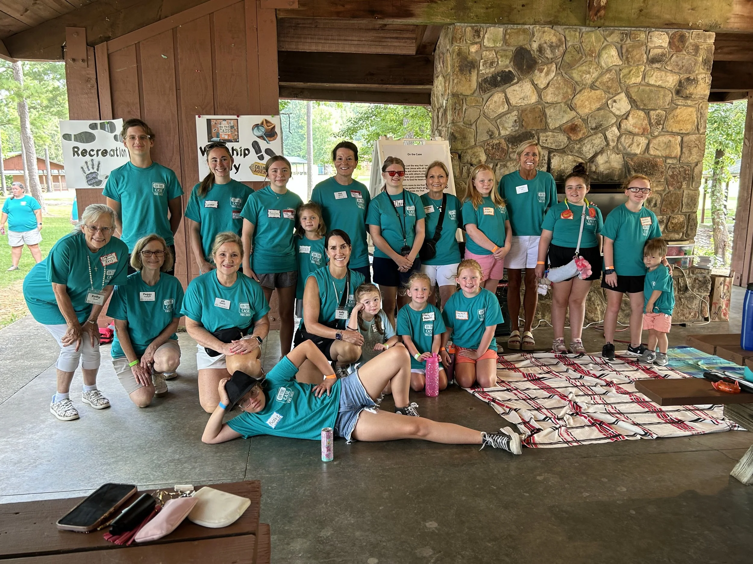A group of children and adults wearing teal shirts posing for a photo at a camp or outdoor event, with a stone fireplace in the background and signs reading 'Recreation' and 'Partnership'.
