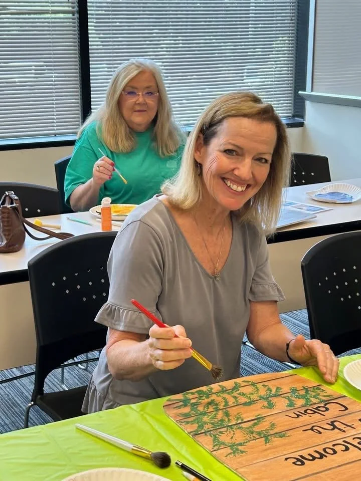 Two women sitting at a table, painting a wooden board with green paint. The woman in the front is smiling, holding a paintbrush, and appears to be painting a decorative design on the board. The woman in the background is also holding a paintbrush and