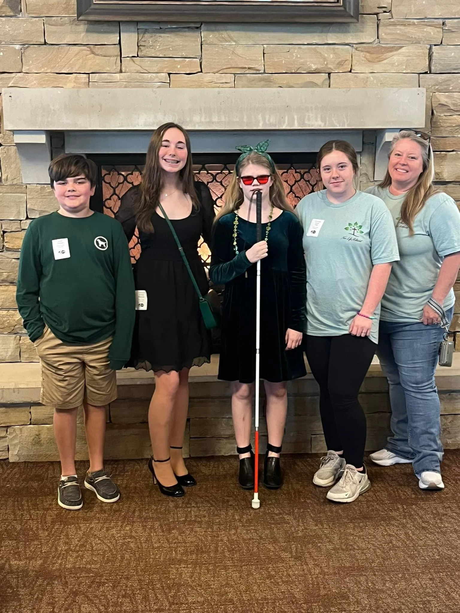 Group of five people, including children and a woman, standing in front of a stone fireplace, with some dressed in casual clothing and one girl wearing sunglasses, a headband, and holding a white cane.