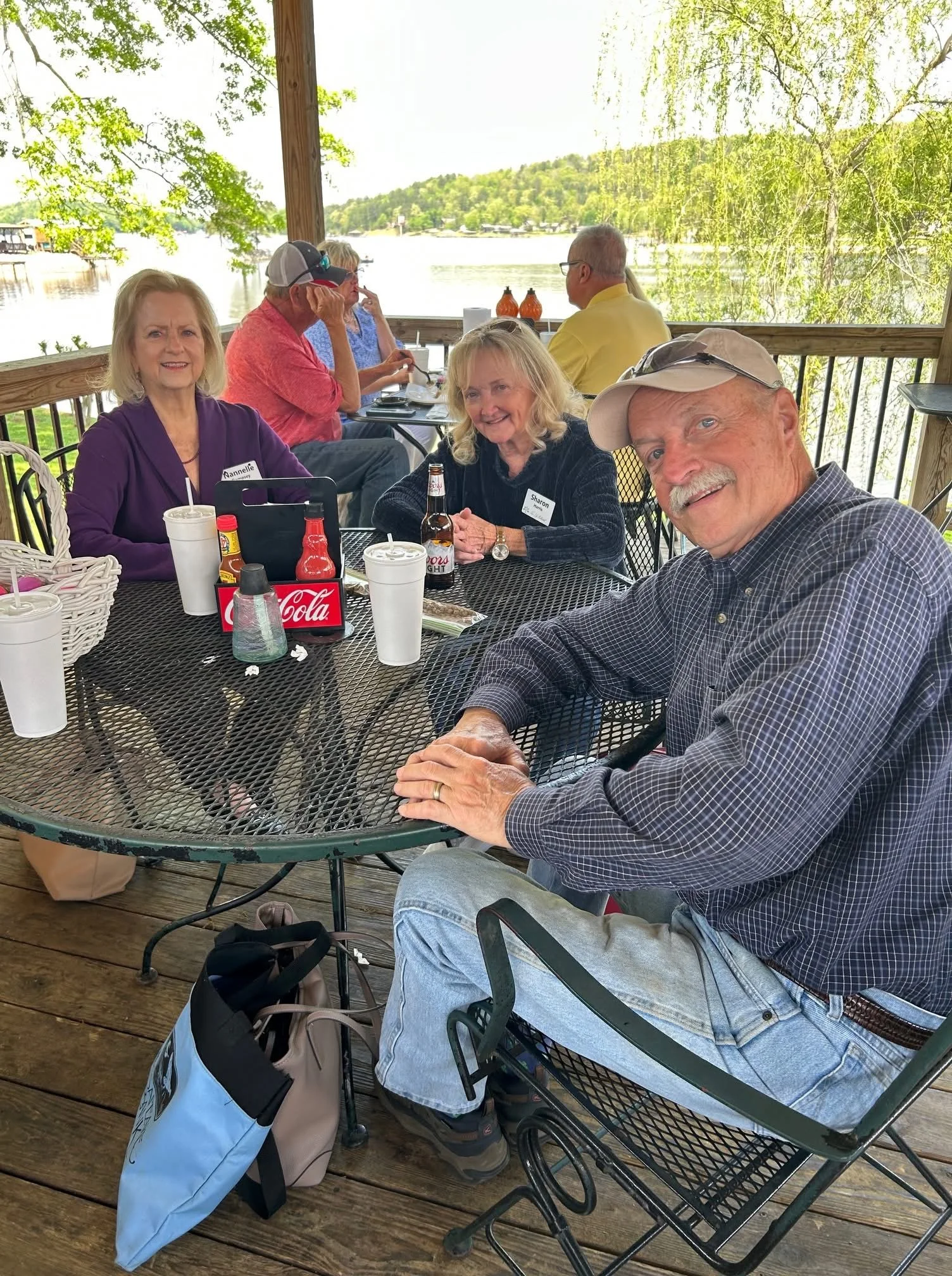 Group of six people sitting at a circular outdoor table near a body of water with trees in the background. Two women and one man are in the foreground smiling at the camera; three other people are in the background, engaged in conversation. The table