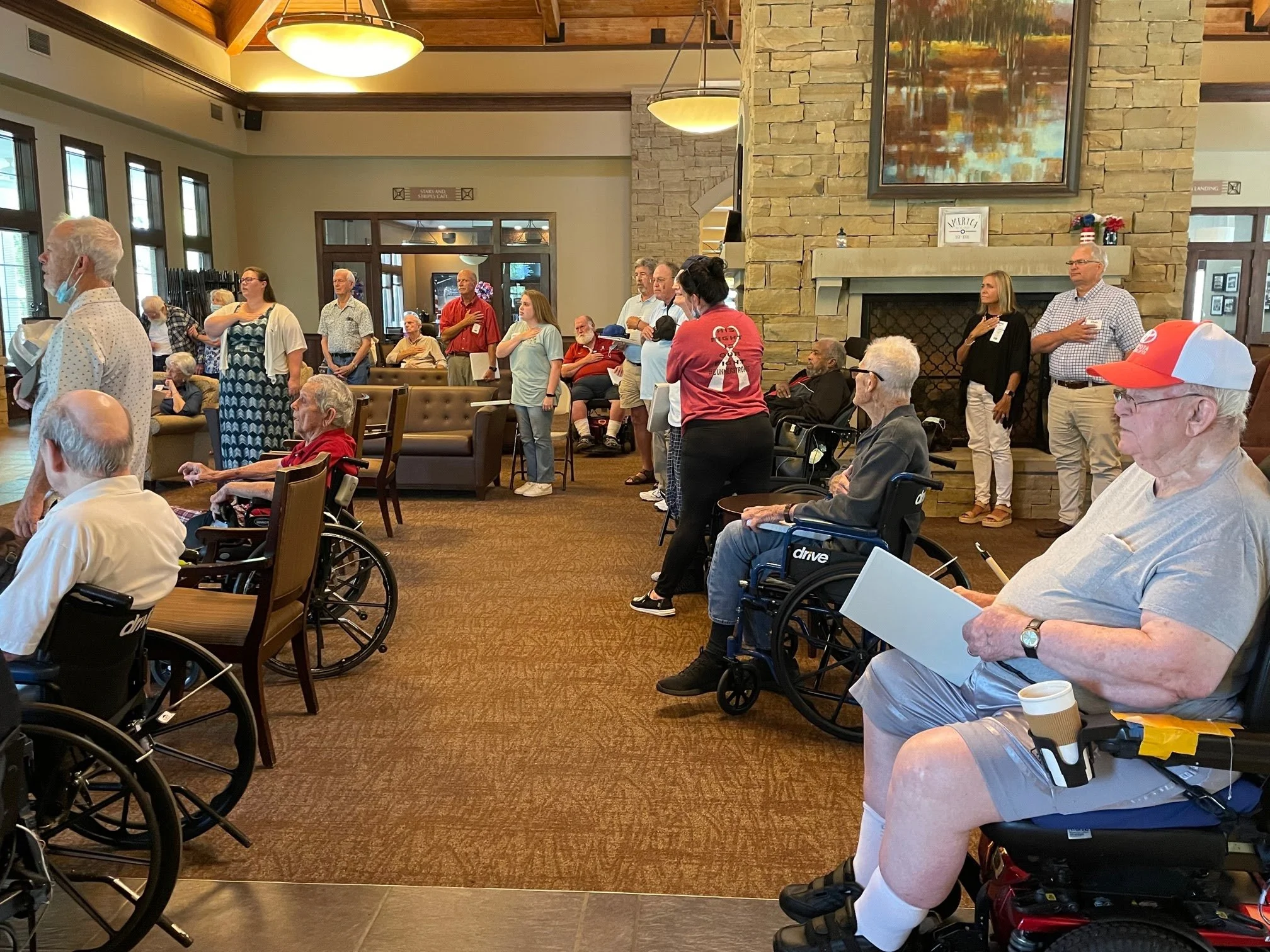 A group of elderly people and caregivers gathered in a spacious room with a fireplace. Some are seated in wheelchairs, some are standing, and others sit on chairs or sofas. Participants are listening and participating in a ceremony or tribute, with s