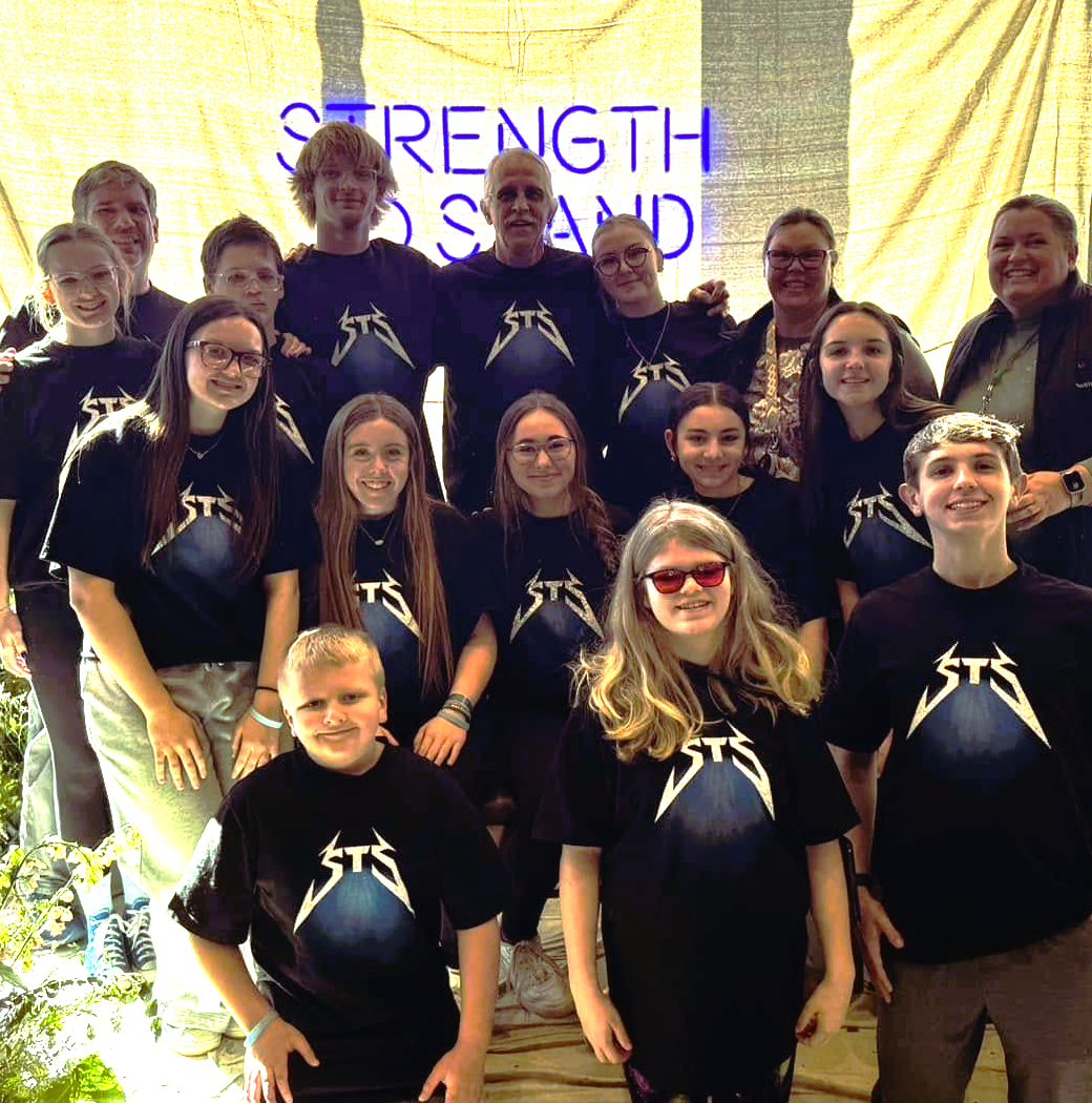 Group of people, mostly young girls and teens, wearing black t-shirts with a rock band logo, standing on a stage with a yellow background and purple neon sign that reads 'Strength of Sound.'