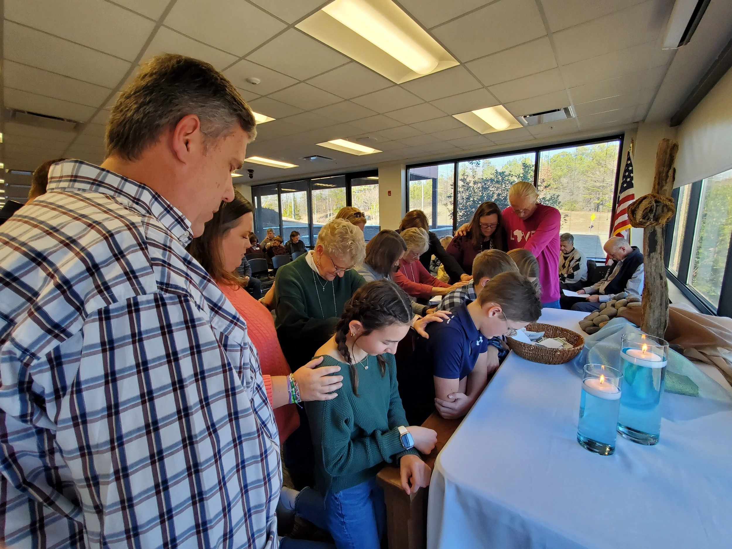 People kneeling and praying around a table during a religious or memorial service in a well-lit room with large windows. There are lit candles on the table.