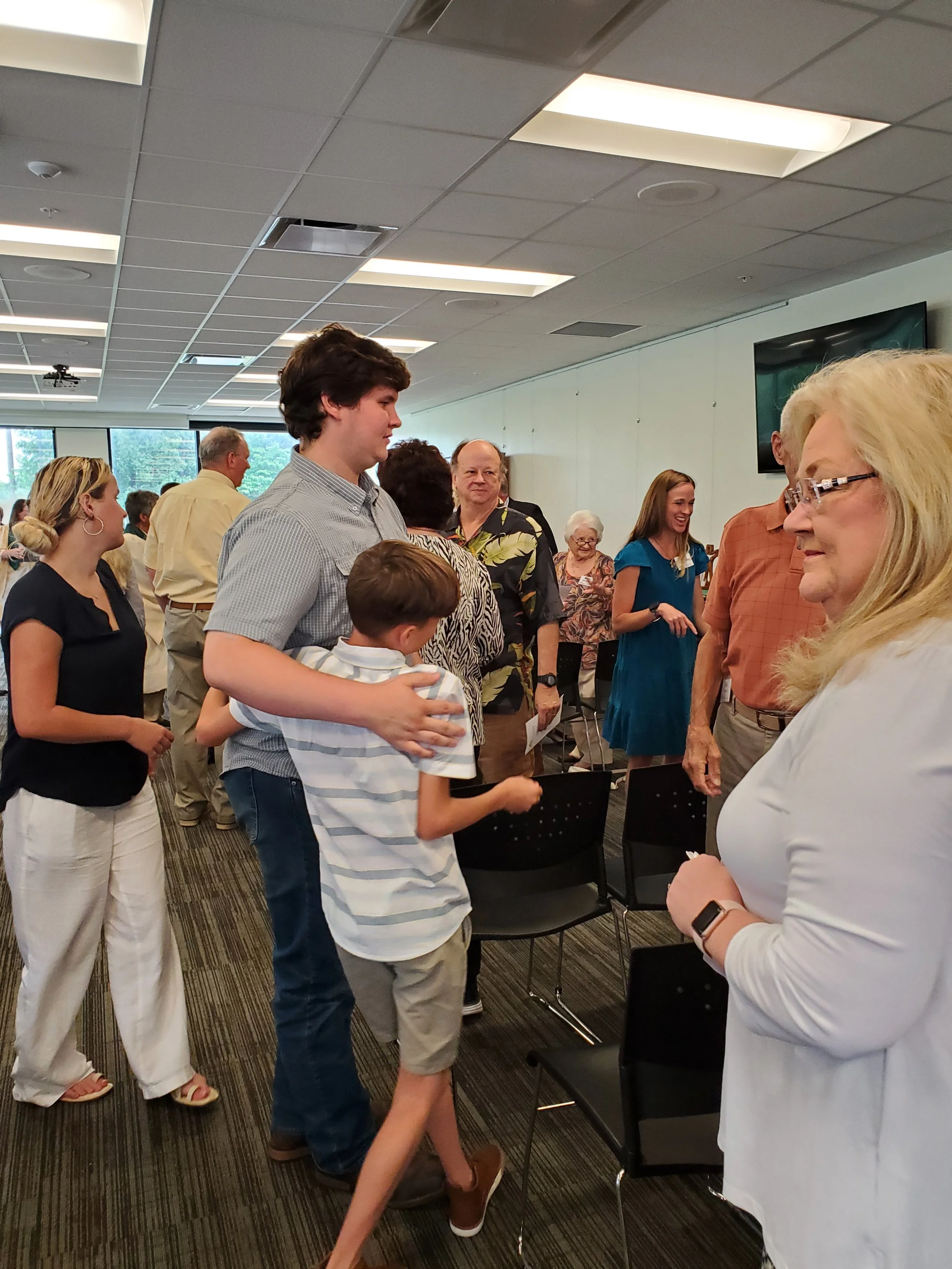 A group of people in a room, some standing and some sitting, engaged in conversation and socializing, with large windows and a television screen in the background.