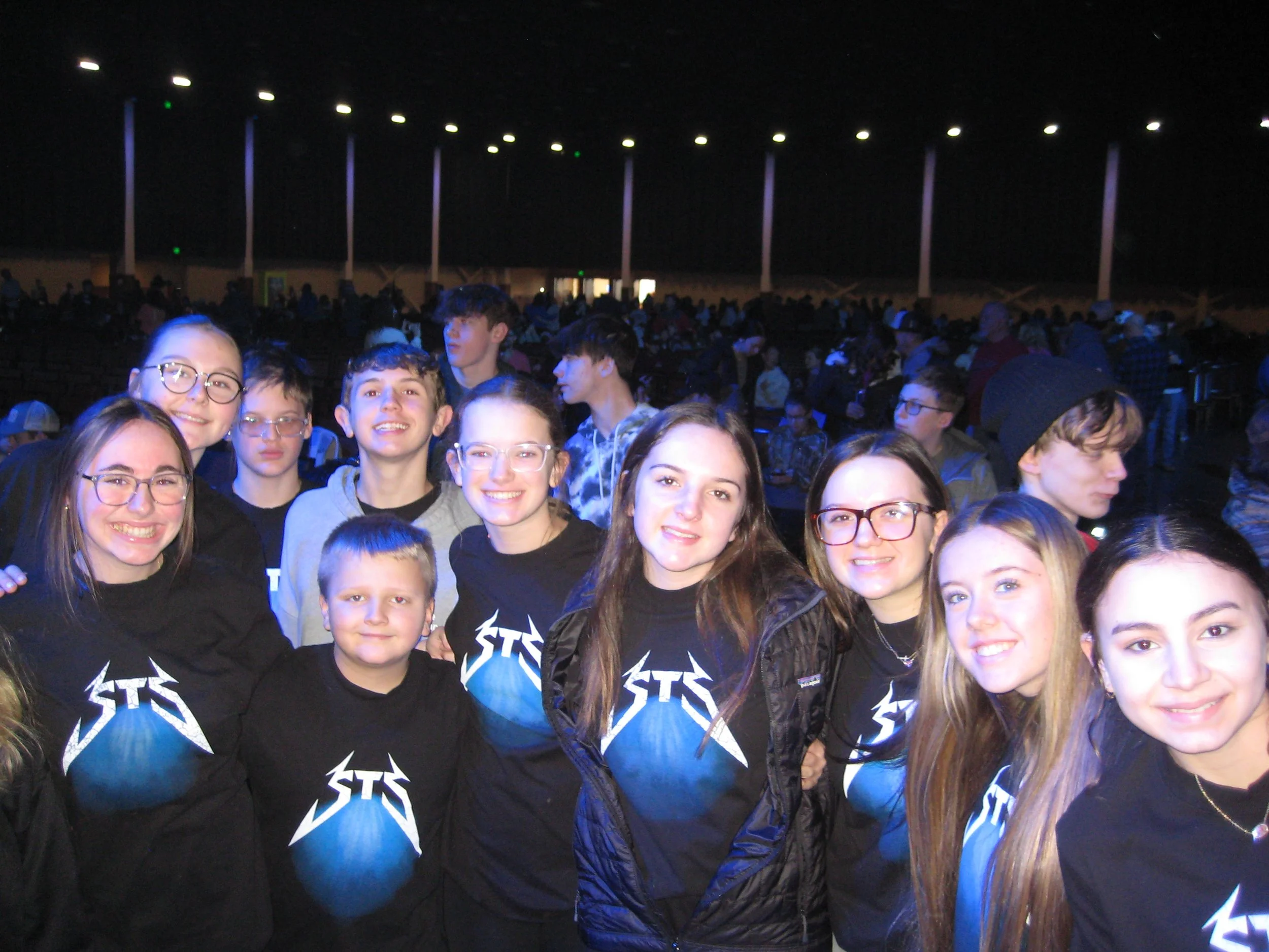 Group of people, mostly young women and children, smiling and posing for the camera at an indoor event, with a dark background, some wearing black T-shirts with a lightning bolt logo.