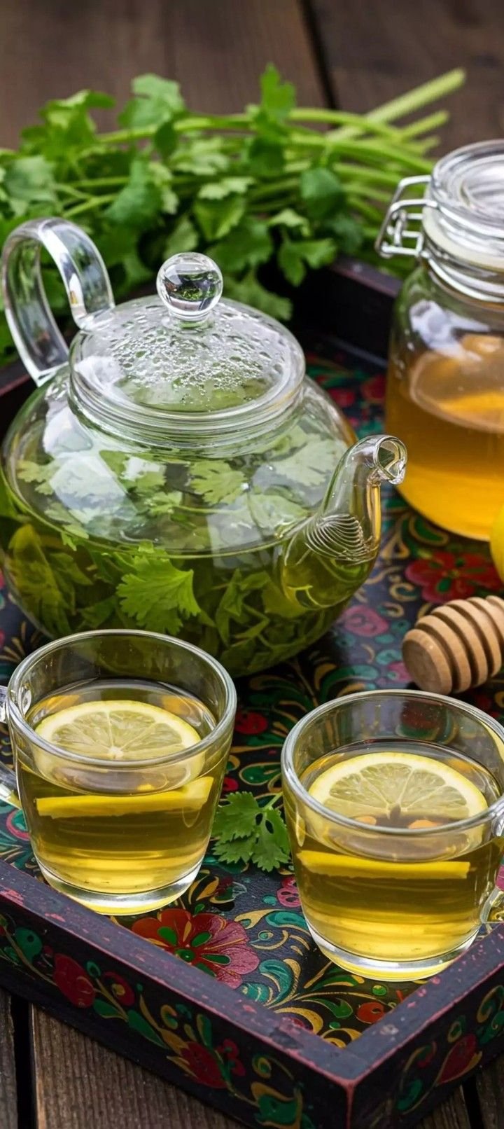 Glass teapot with herbs and water, two glasses of herbal tea with lemon slices, a jar of honey, a honey dipper, fresh cilantro, and a colorful tray on a wooden surface.