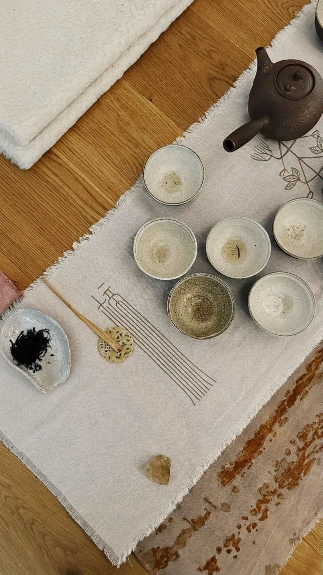 Tea set with six tea cups, a teapot, a small dish with tea leaves, and decorative objects on a white table runner on a wooden floor.