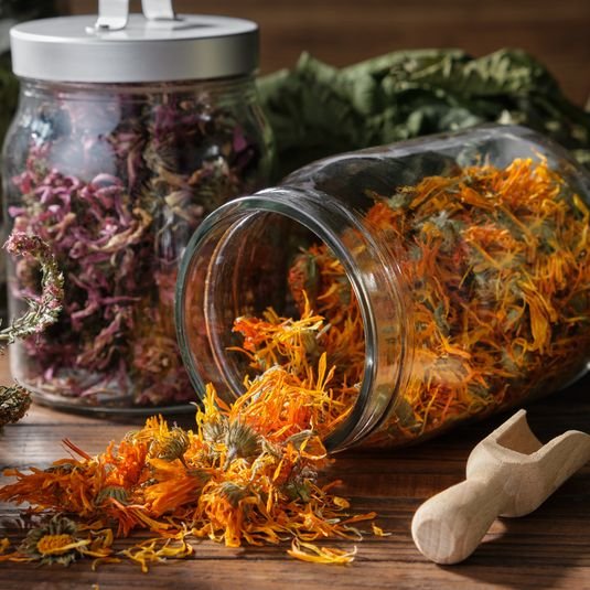 Glass jar filled with dried orange calendula flowers, with some flowers spilled out on a wooden surface. In the background, there is another jar filled with dried purple flowers and some green leaves.