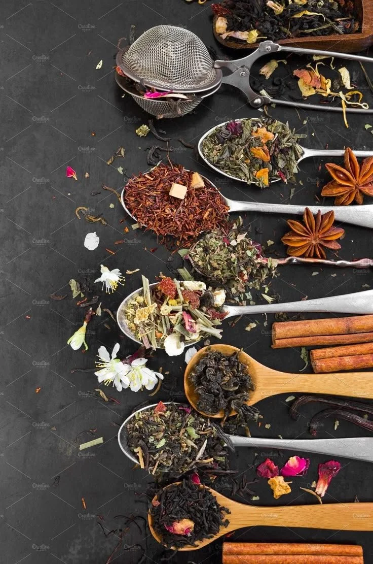 Various loose dried herbs, flowers, and spices arranged on spoons and a black surface, some with star anise and cinnamon sticks.