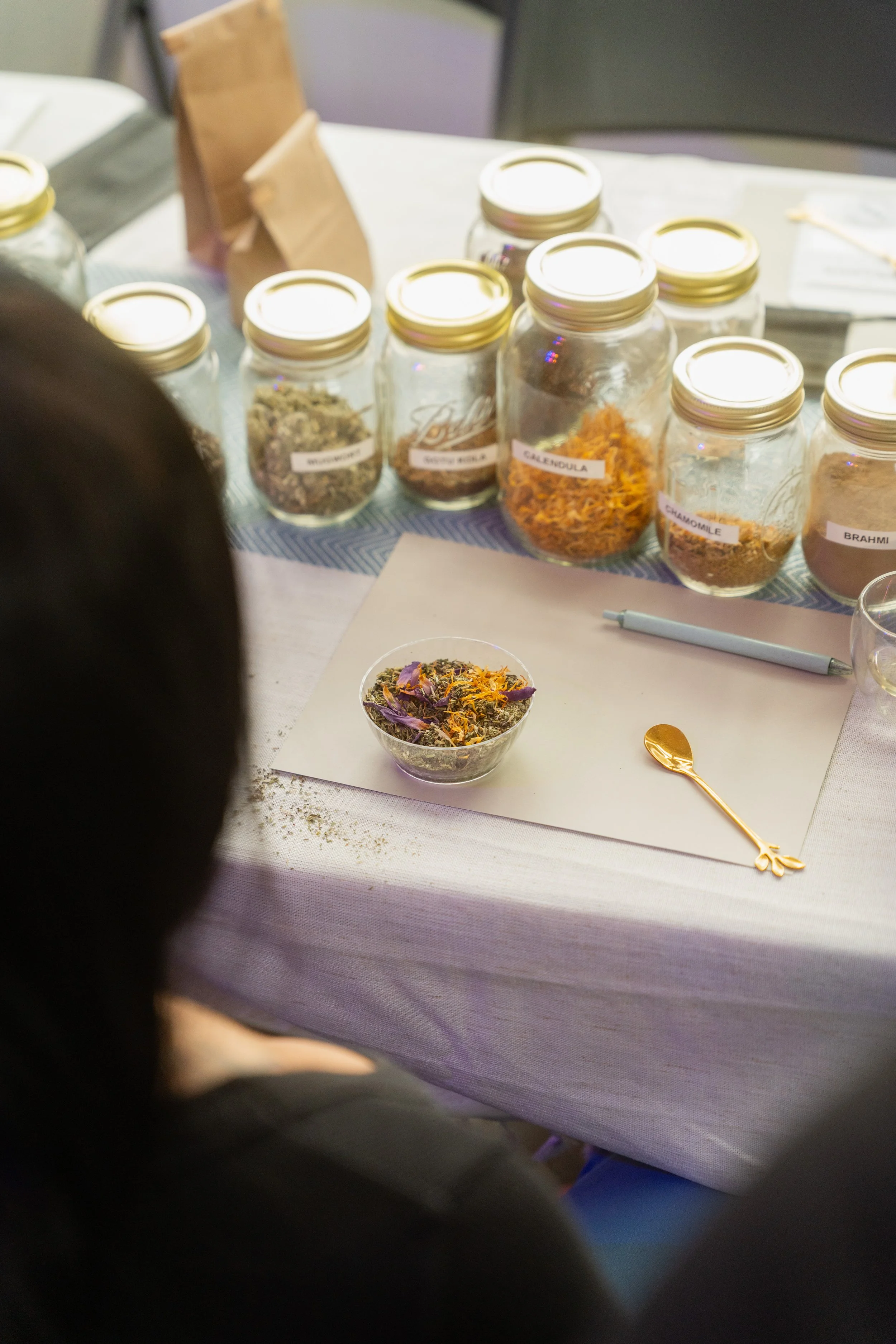 Display of various labeled jars of dried herbs and flowers on a table, with a small bowl of mixed herbs and a gold spoon in front.