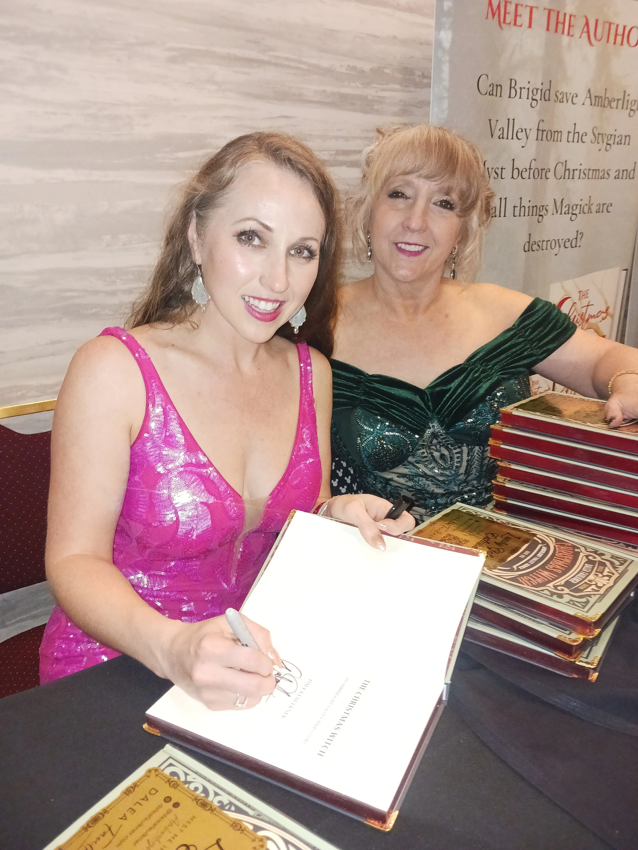 Dalea Faulkner Author and Martina webster Producer Two women at a book signing event, one in a pink dress signing a book, the other in a green dress smiling, with stacks of books on the table.