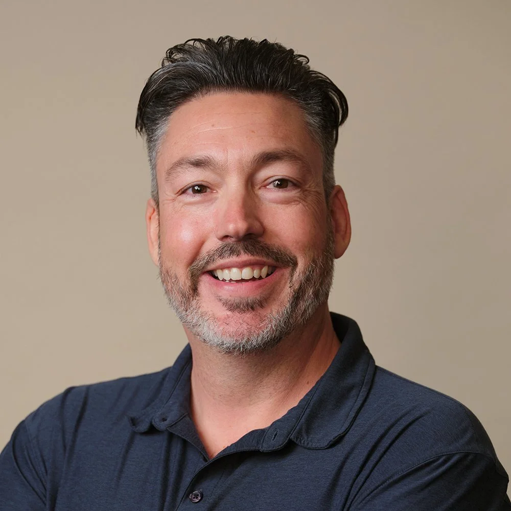 A smiling middle-aged man with dark hair, a beard with gray, wearing a navy polo shirt, against a beige background.