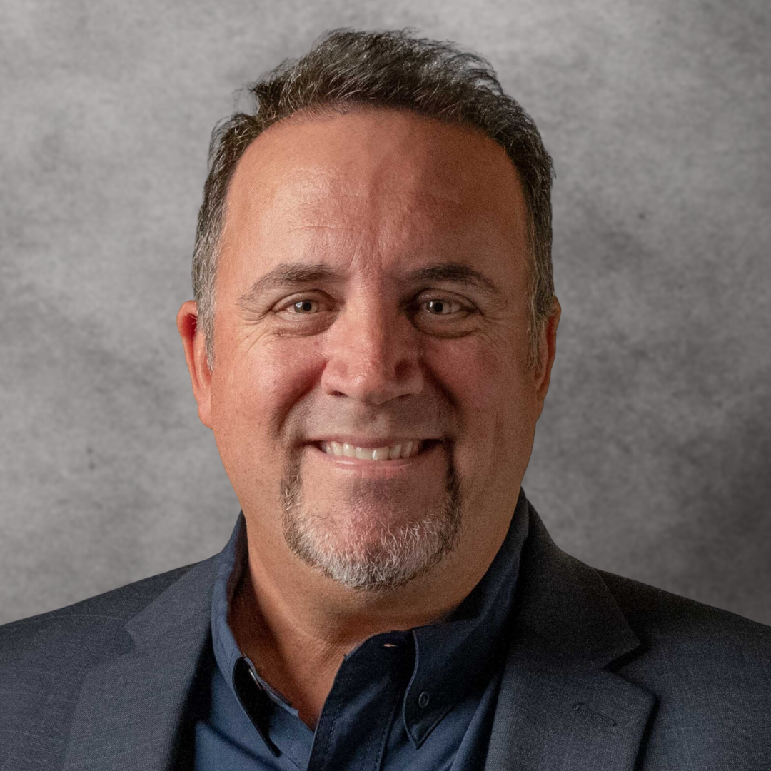A middle-aged man with short dark hair, sporting a beard and mustache, smiling and wearing a dark blazer and collared shirt, against a gray background.
