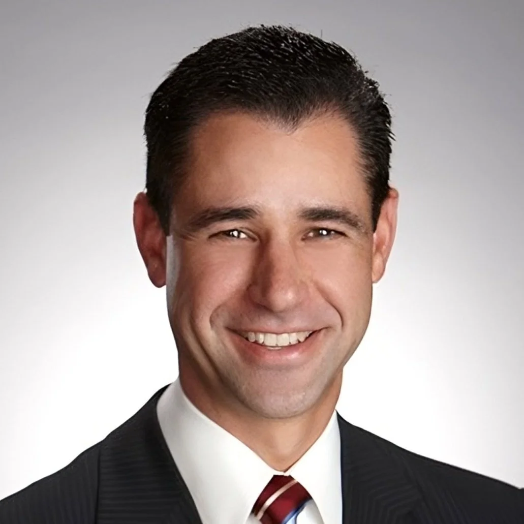 A professional headshot of a smiling man in a suit and tie, with dark hair, against a gray gradient background.
