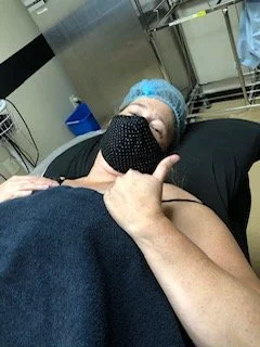 Patient in hospital bed wearing a face mask and a surgical cap giving a thumbs-up gesture.