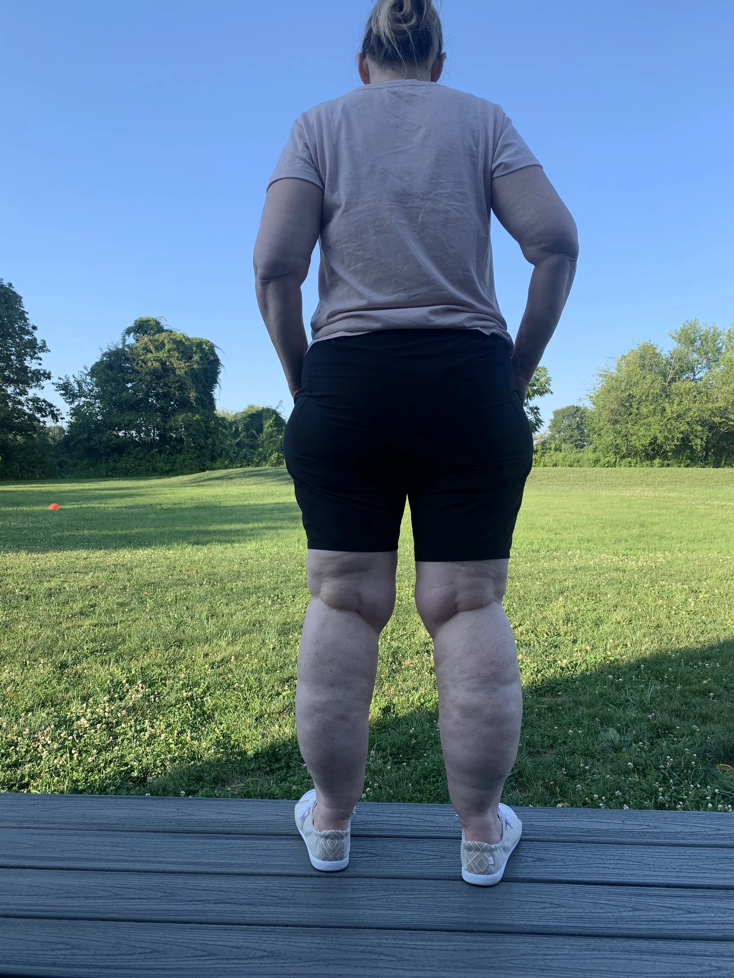 Person standing on a deck overlooking a grassy field, wearing a pink shirt, black shorts, and white shoes, with their back facing the camera.