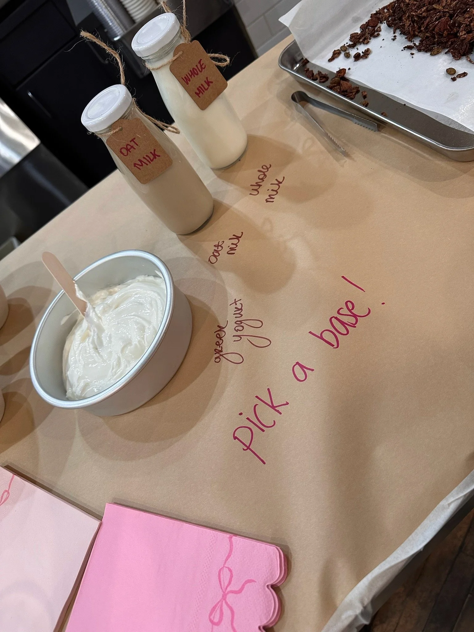 A table setup for making yogurt with bottles of oat and whole milk, a bowl of plain yogurt with a spoon, a tray of chocolate chips, pink napkins, and a motivational message written on the table that says, 'pick a base!'
