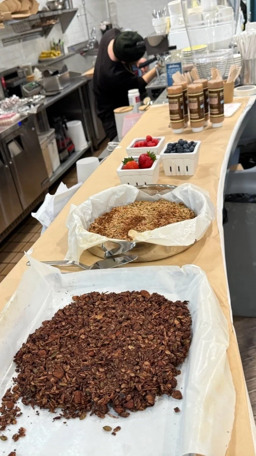 Breakfast bar with granola, fresh strawberries, blueberries, and various coffee drinks in cups, with a barista preparing coffee in the background.